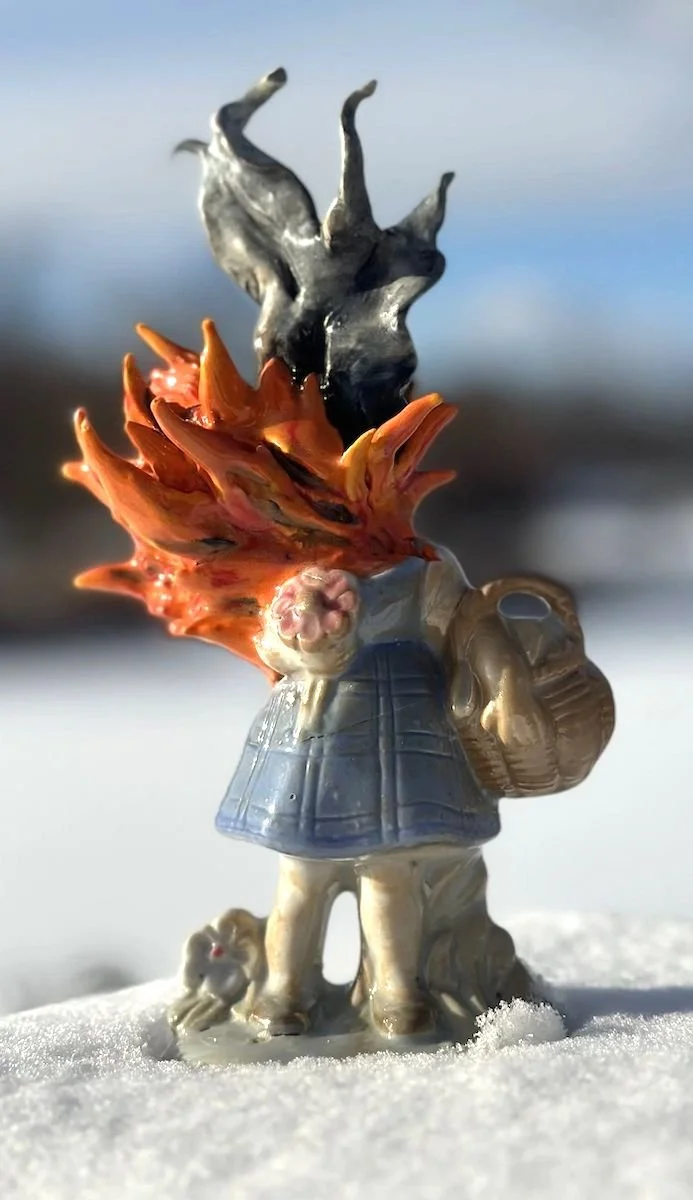 A figurine of a girl in a coat standing in the snow, with a dog at her side, and an flames/explosion coming from her head, against a background of a snowy landscape and blue sky.