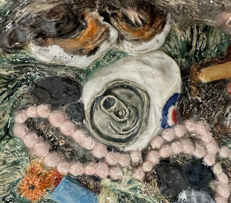 Close-up of a collection of trash including a trompe l'oeil crushed soda can, plastic beads, a piece of orange sponge, and other debris submerged in water.