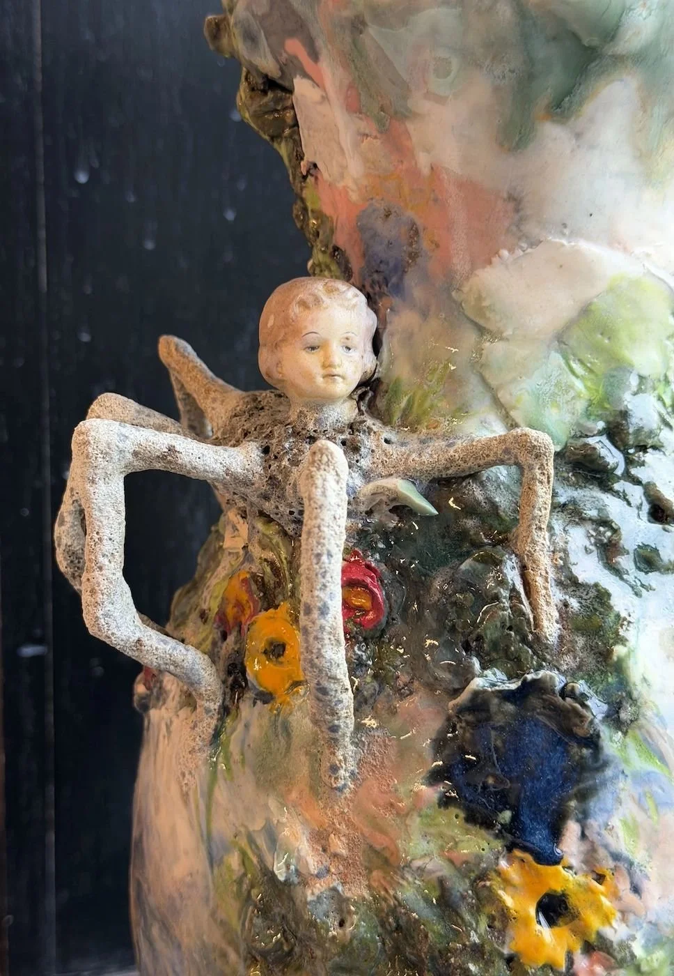 A mixed media sculpture featuring a child's face attached to an organic textured structure with flowing, colorful glazes and abstract forms.