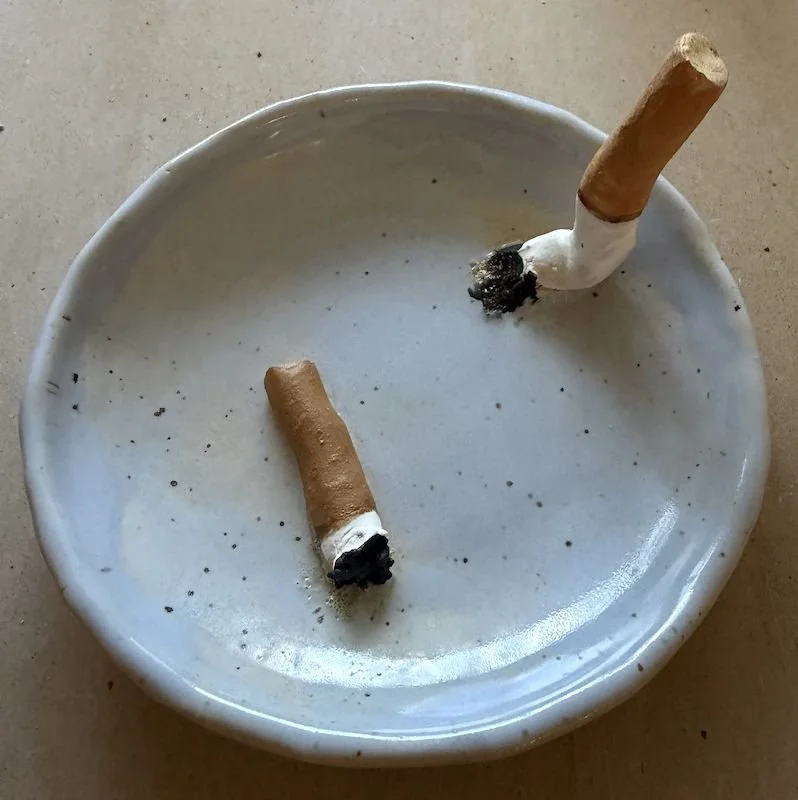 Empty ceramic ashtray with two trompe l'oeil ceramic cigarettes, one resting on the edge and the other lying inside, both burned out.