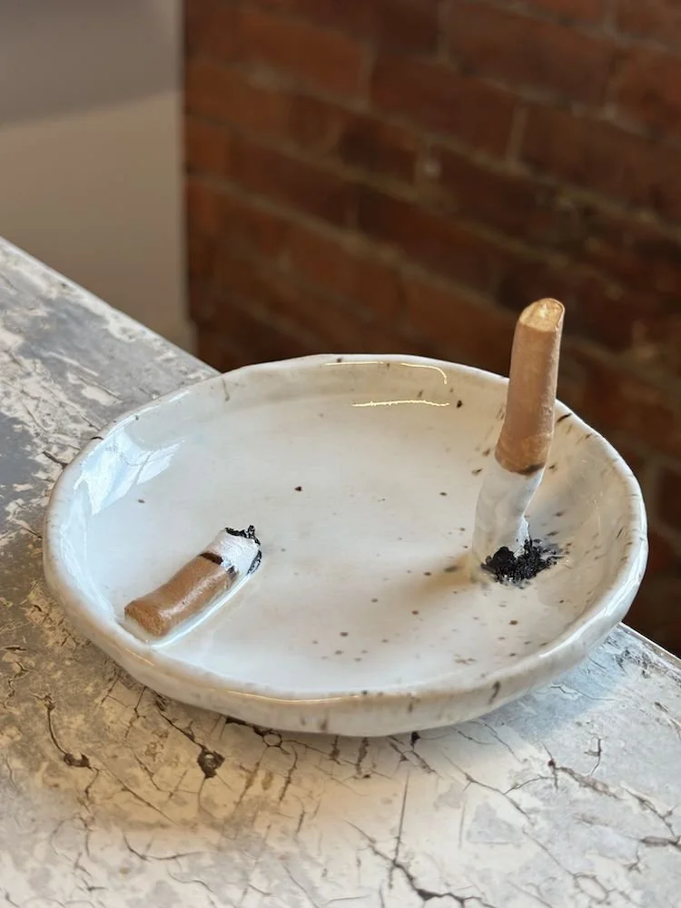 A ceramic ashtray with two cigarette butts, one partially burned and the other more intact, placed on a distressed white surface against a brick wall background.