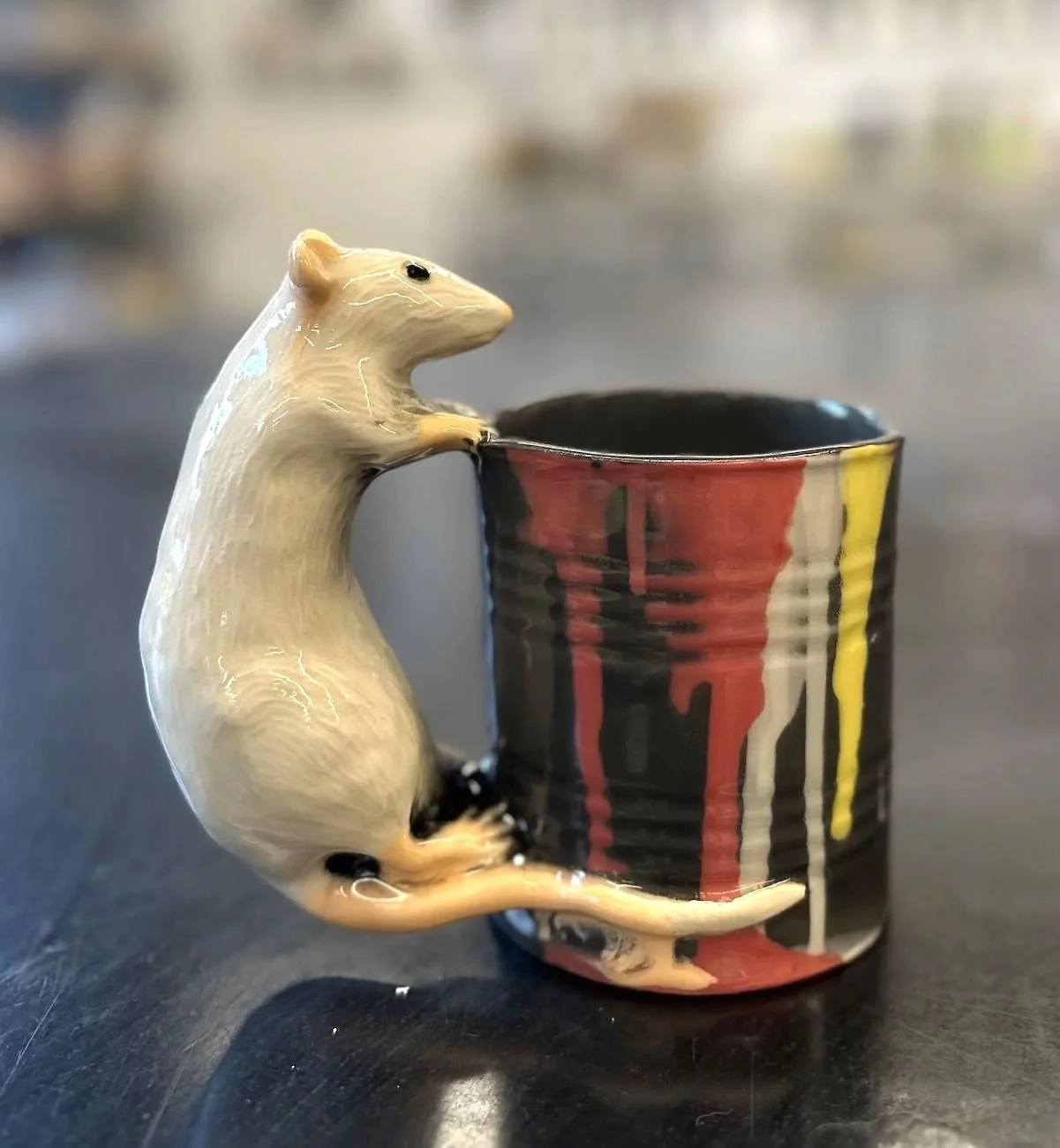A ceramic mug with red, yellow, and white drips on a black background, featuring a beige mouse-shaped handle.