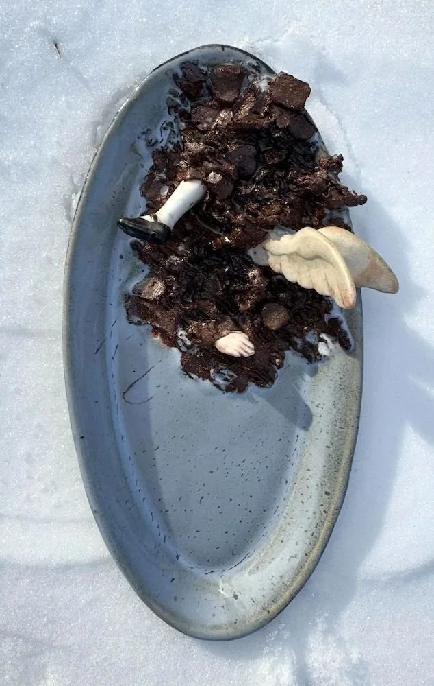 A silver dish with dark, melted chocolate and a white chocolate angel wing on top, placed on snow.