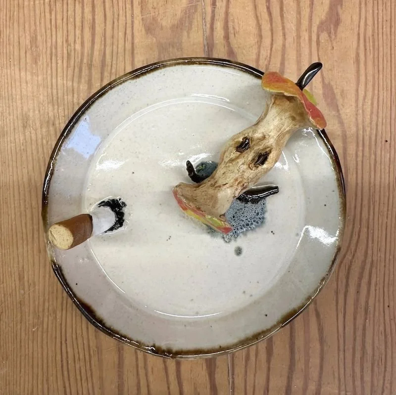 Empty ashtray with a large mushroom-shaped object and a small cigarette butt, on a wooden surface.