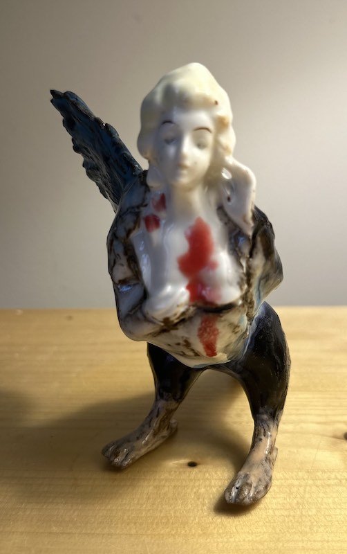 A ceramic figurine of a humanoid dog with a human face, holding its right hand to its ear. The figurine has black wings on its back and appears to be distressed, with red markings on its chest.