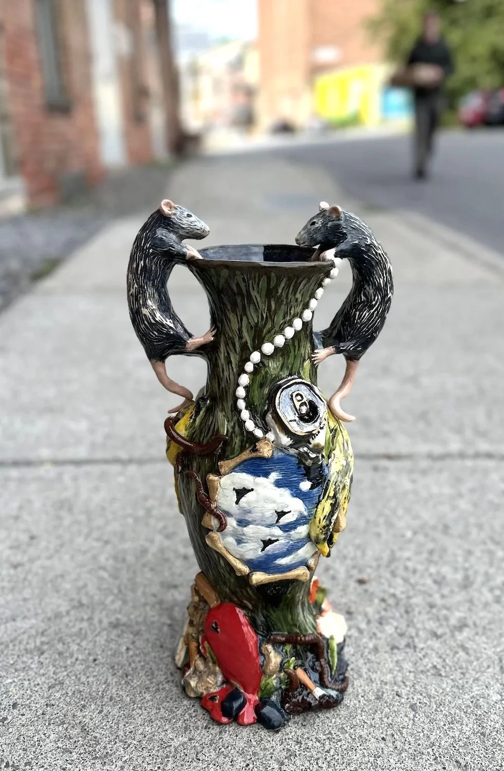Decorative ceramic vase with two black and white rats on the sides, featuring other details like a skull, a red object, and a cloudy sky painted on it, set on a sidewalk outdoors.