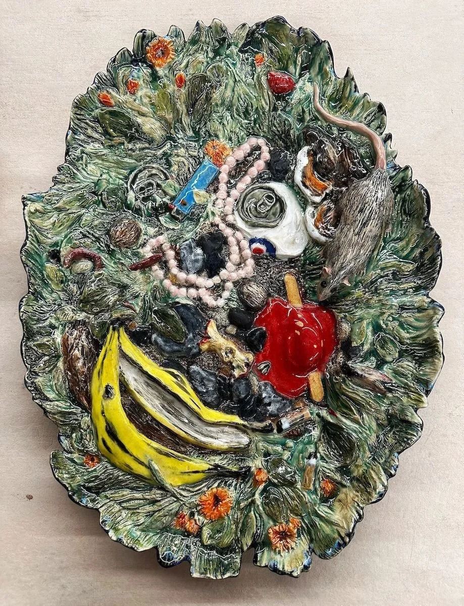 Colorful ceramic dish resembling a leafy platter filled with various miniature objects including a banana, skull, mouse, red popcicle, beads, and small figurines.