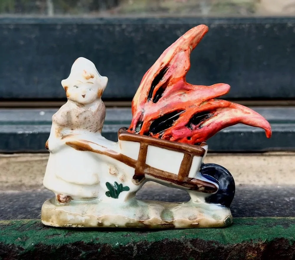 A porcelain figurine of a girl with a ponytail, standing next to a wheelbarrow with a large, fiery red dragon head inside.