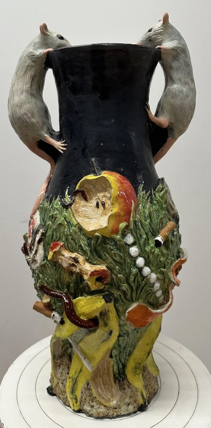 A ceramic sculpture featuring two rats holding a black pot, with a colorful apple, fish, and other food items depicted on the base of the sculpture.