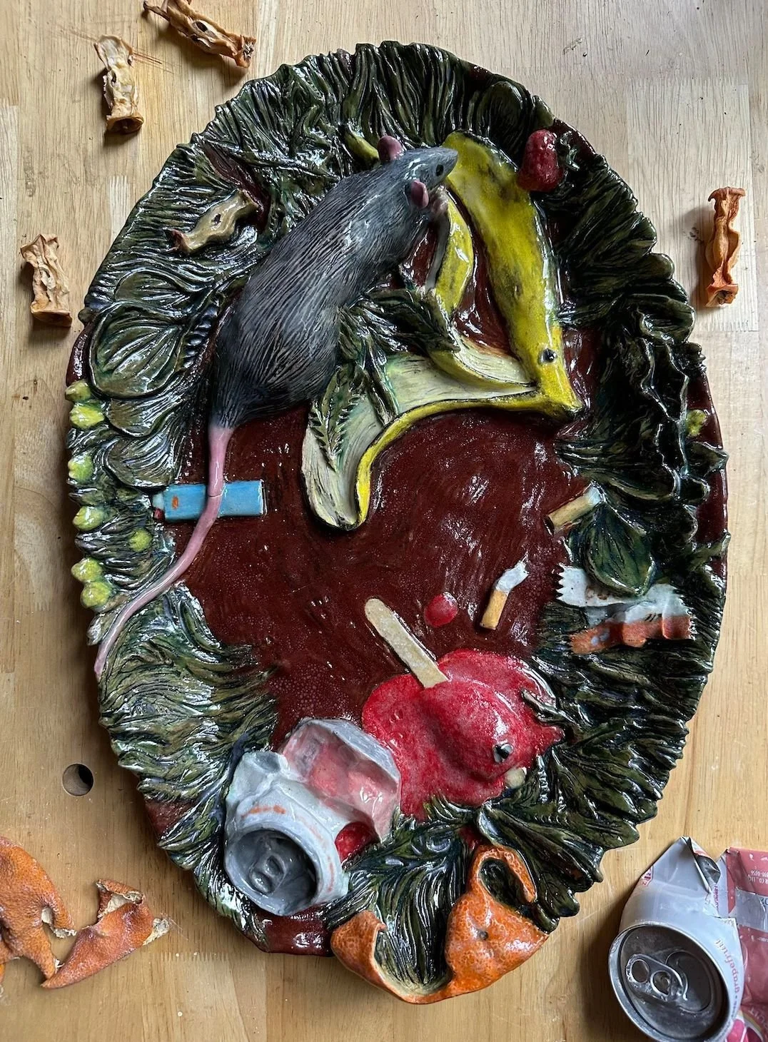 Colorful ceramic or clay sculpture depicting a rat, banana peel, apple, and cans on a leafy background.