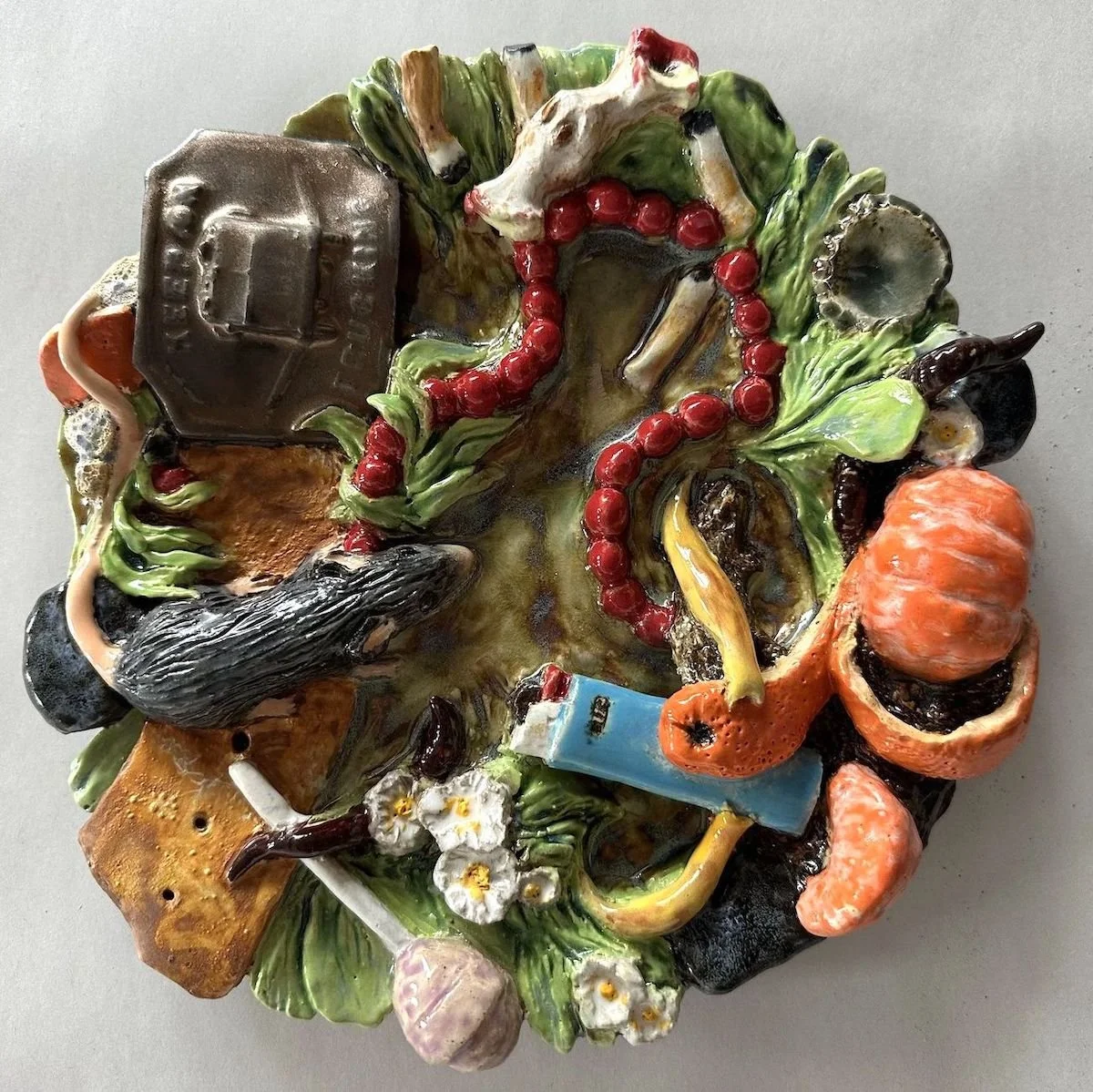 Colorful ceramic sculpture featuring a belt buckle, a string of beads, a lighter, a lollypop, and a mouse