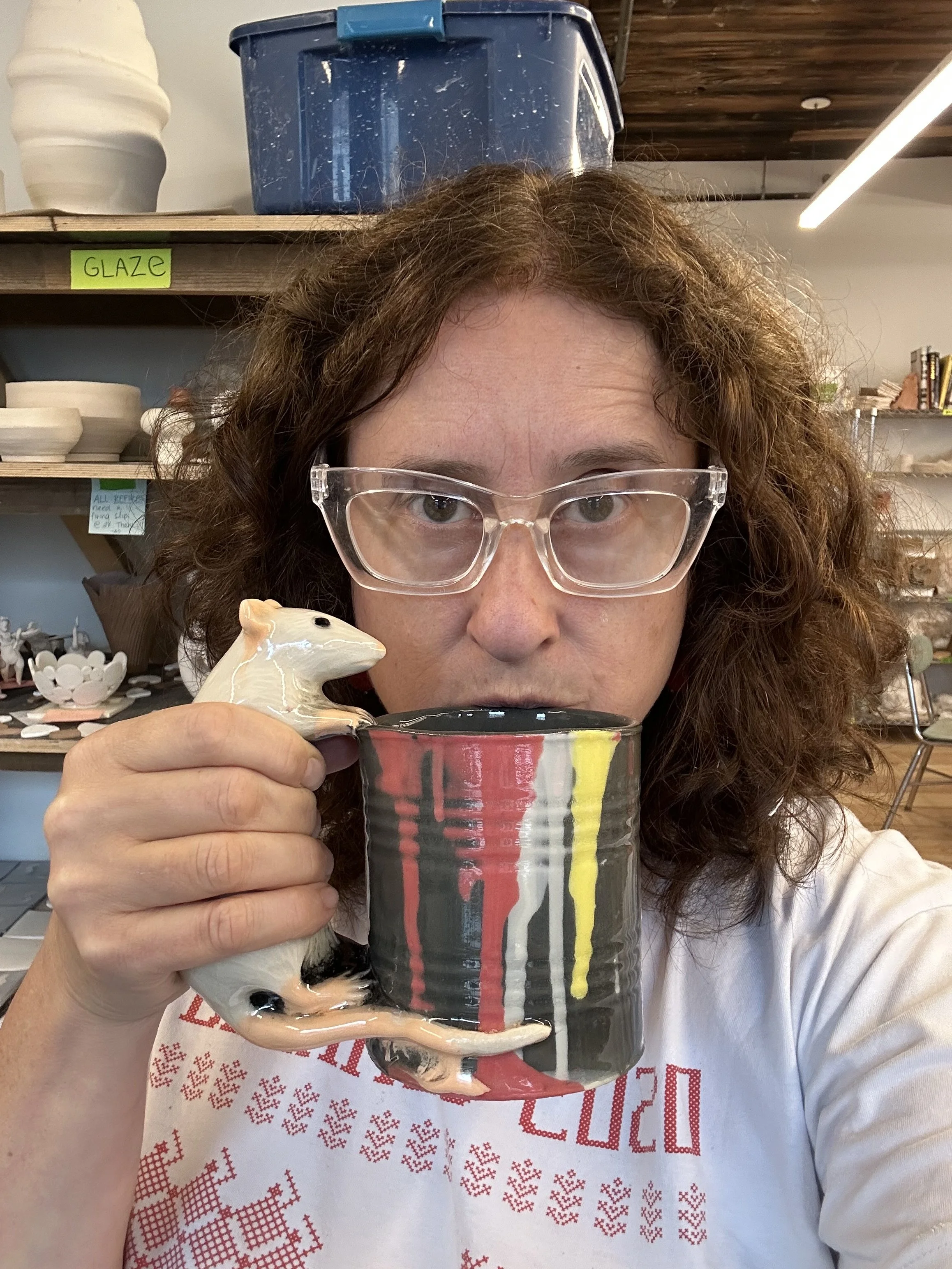 A woman with curly brown hair, wearing clear glasses, holding a ceramic mug with a handle shaped like a rat. Behind her on the shelves are various bowls and containers, including a large white vase, a blue container labeled 'GLAZE'