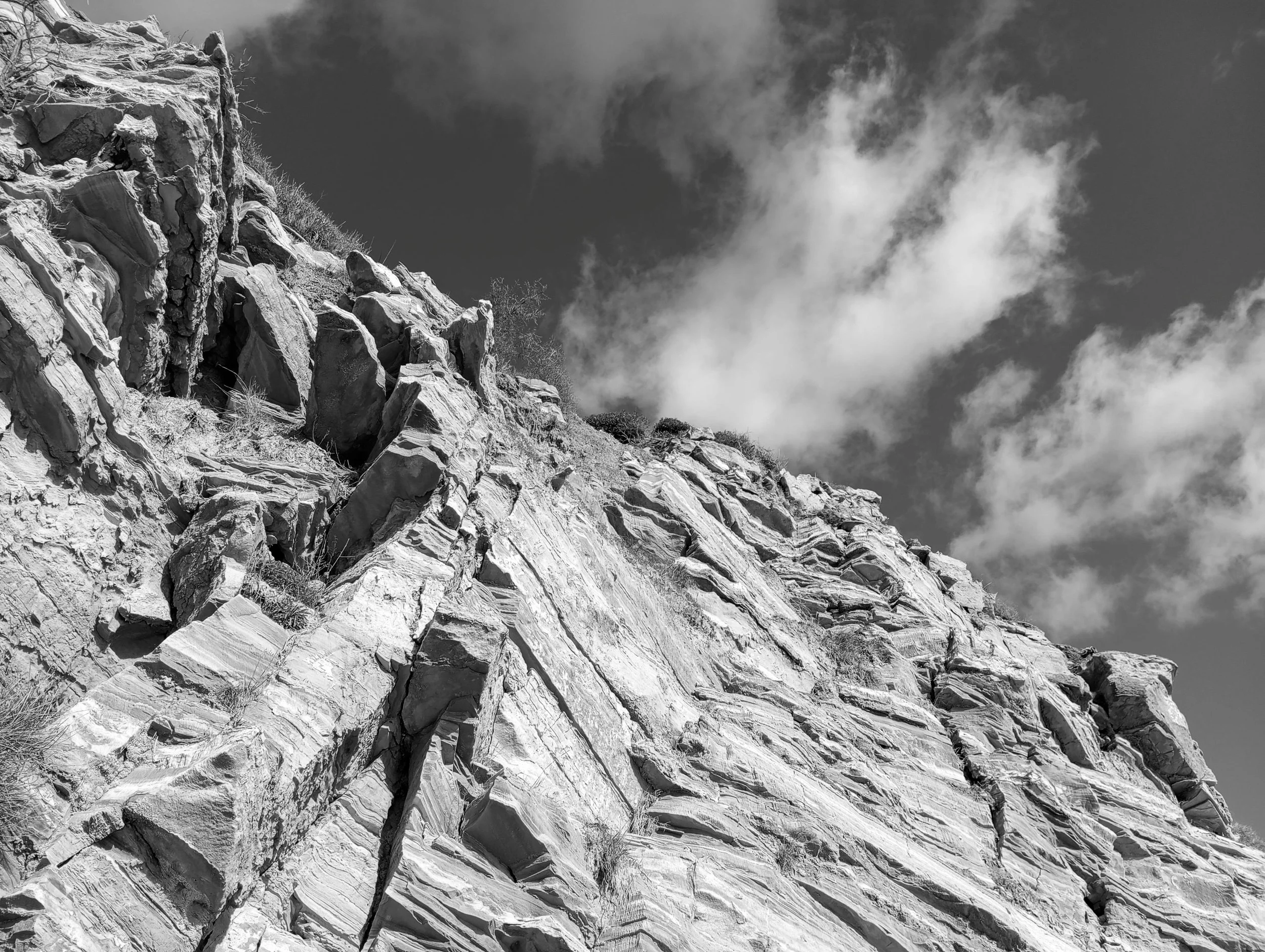 Black and white photograph of a rugged rock cliff taken from a low angle, with sharp, layered formations leading upward toward a partly cloudy sky.