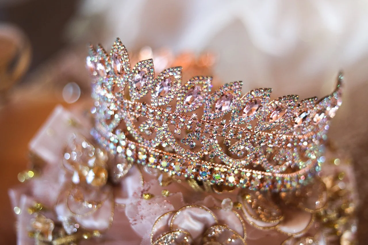A sparkling tiara adorned with pink gemstones and rhinestones, resting on a pink satin surface with golden decorative elements.