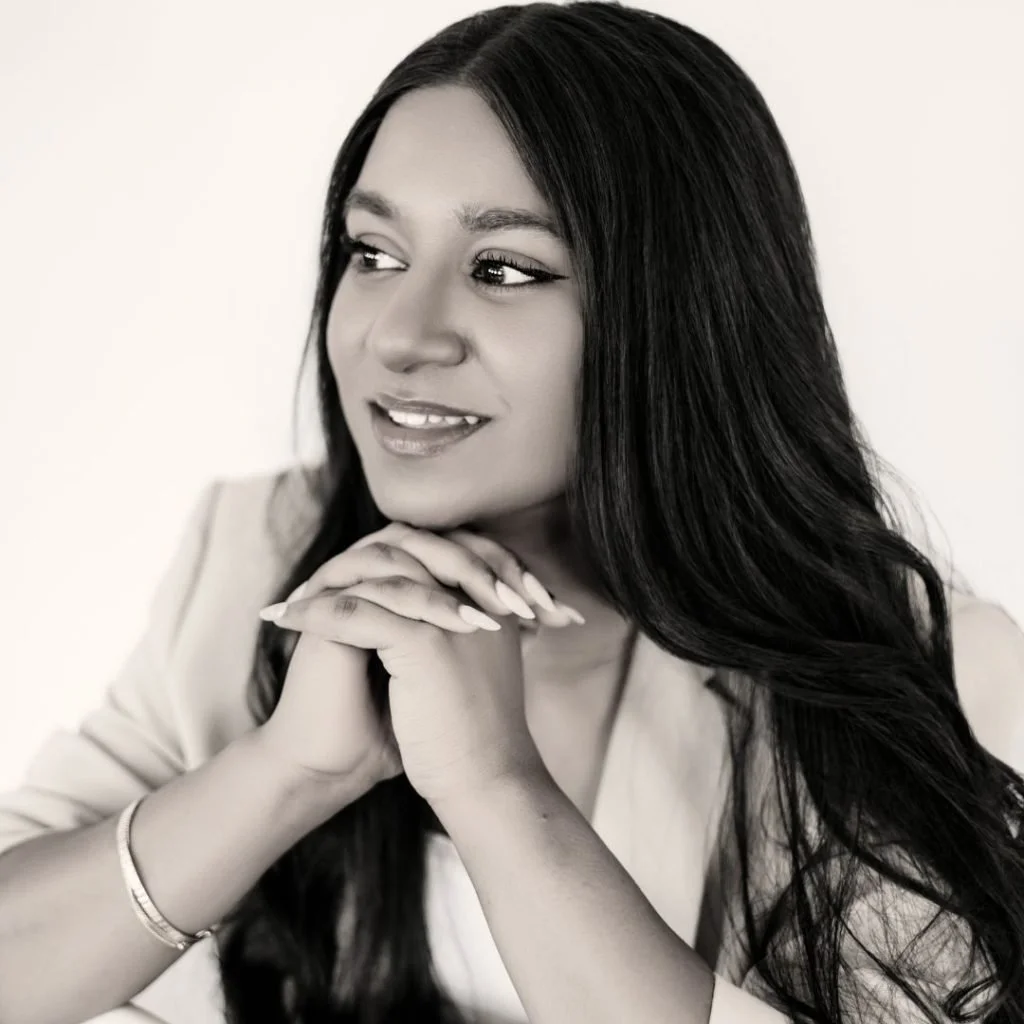 A woman with long hair, posing against a plain wall in black and white, wearing a blazer, a dark crop top, and high-waisted paper bag pants with a bow tie.
