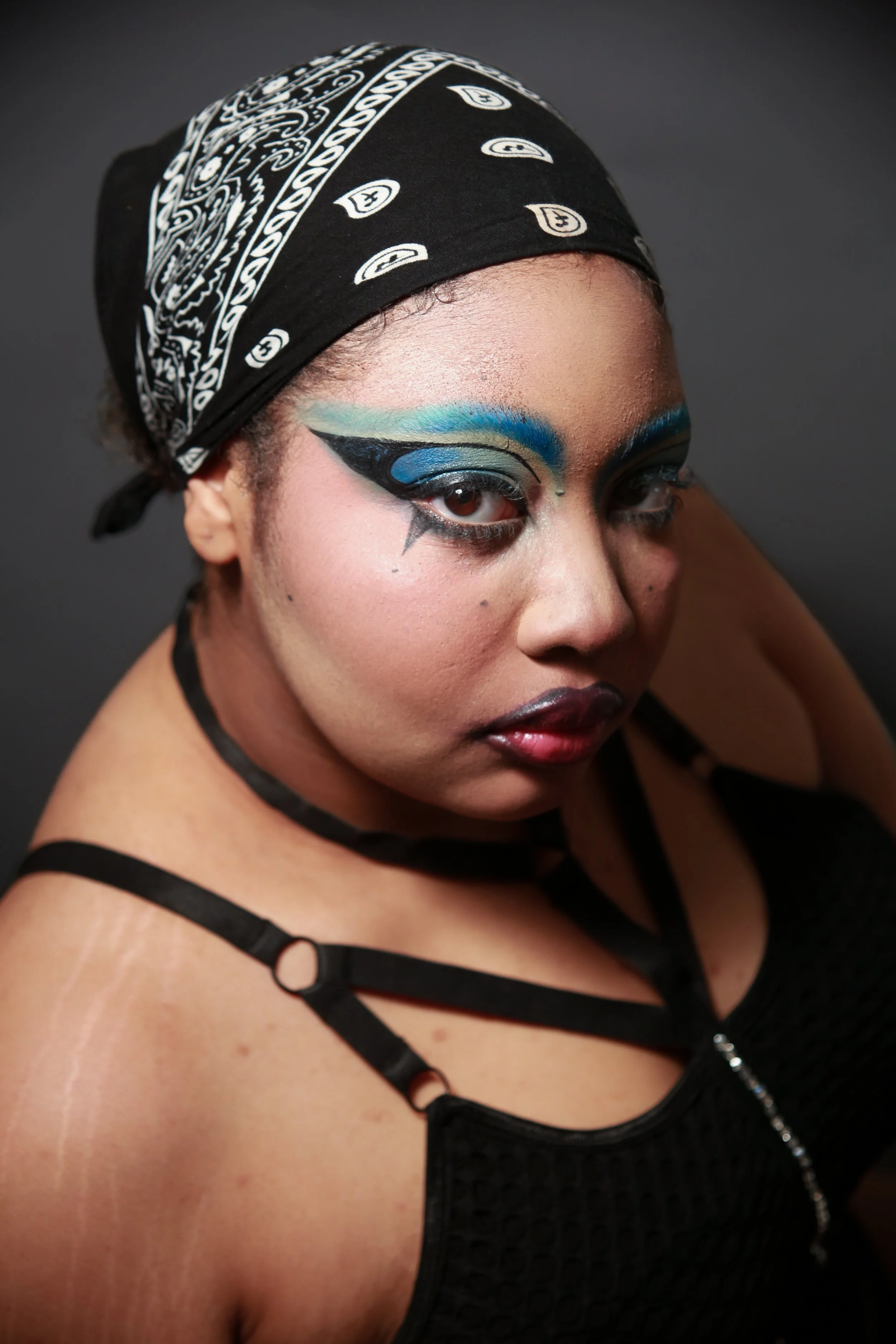 A woman with bold, colorful eye makeup and a black bandana, looking at the camera against a dark background.