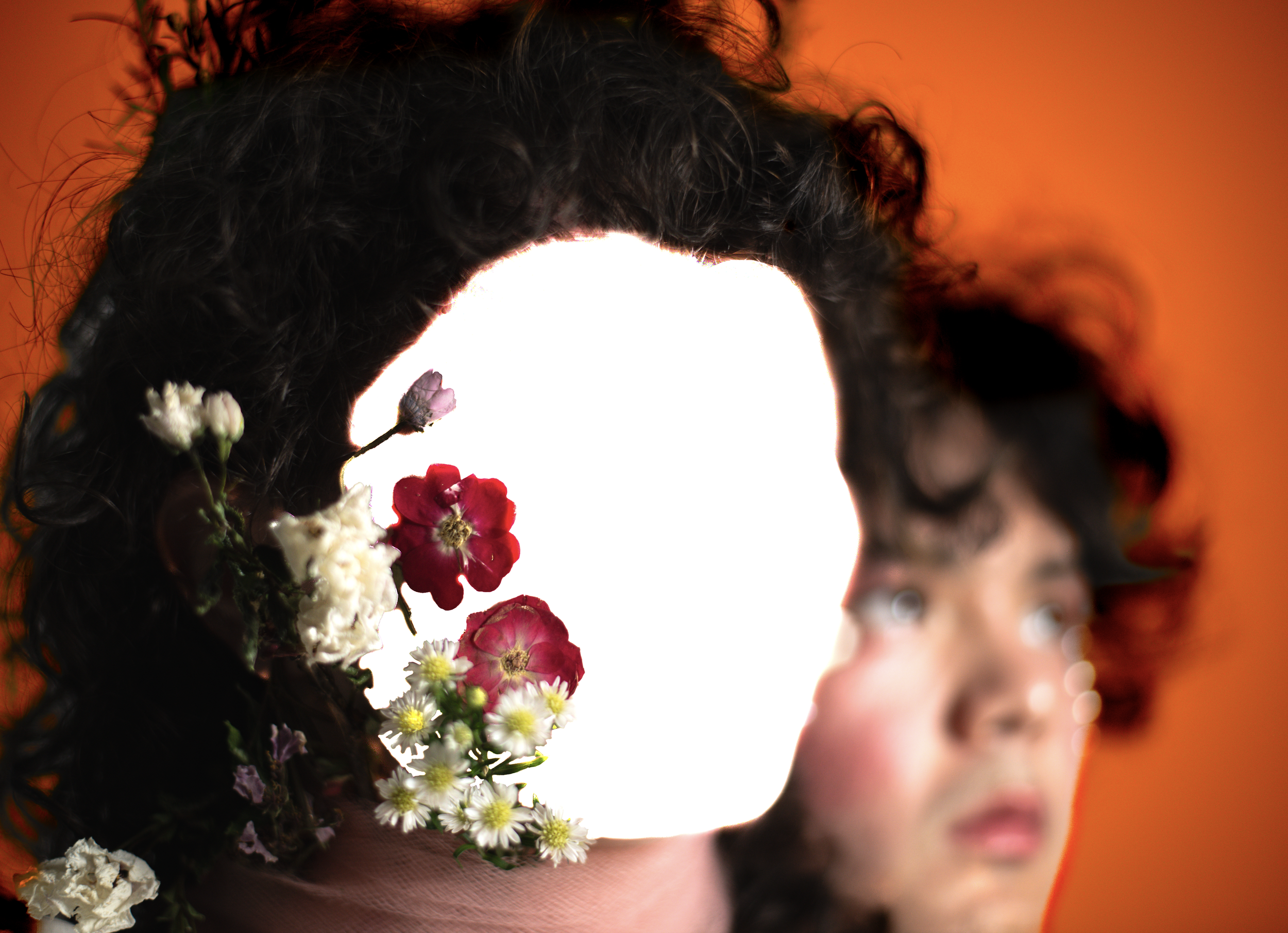 A person with curly dark hair, adorned with various flowers, holding a young child with curly dark hair against an orange background.