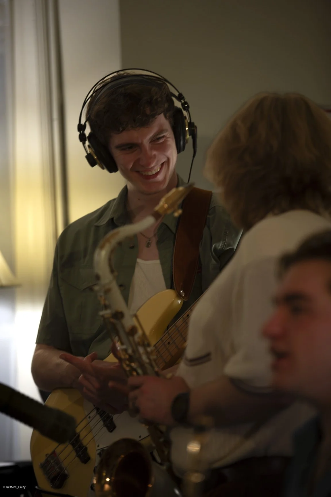 A young man with curly hair wearing headphones and a green shirt, smiling while holding a bass guitar, talking to a blonde woman with a white shirt. There is also a young man with glasses and dark hair, partially visible, in the foreground.