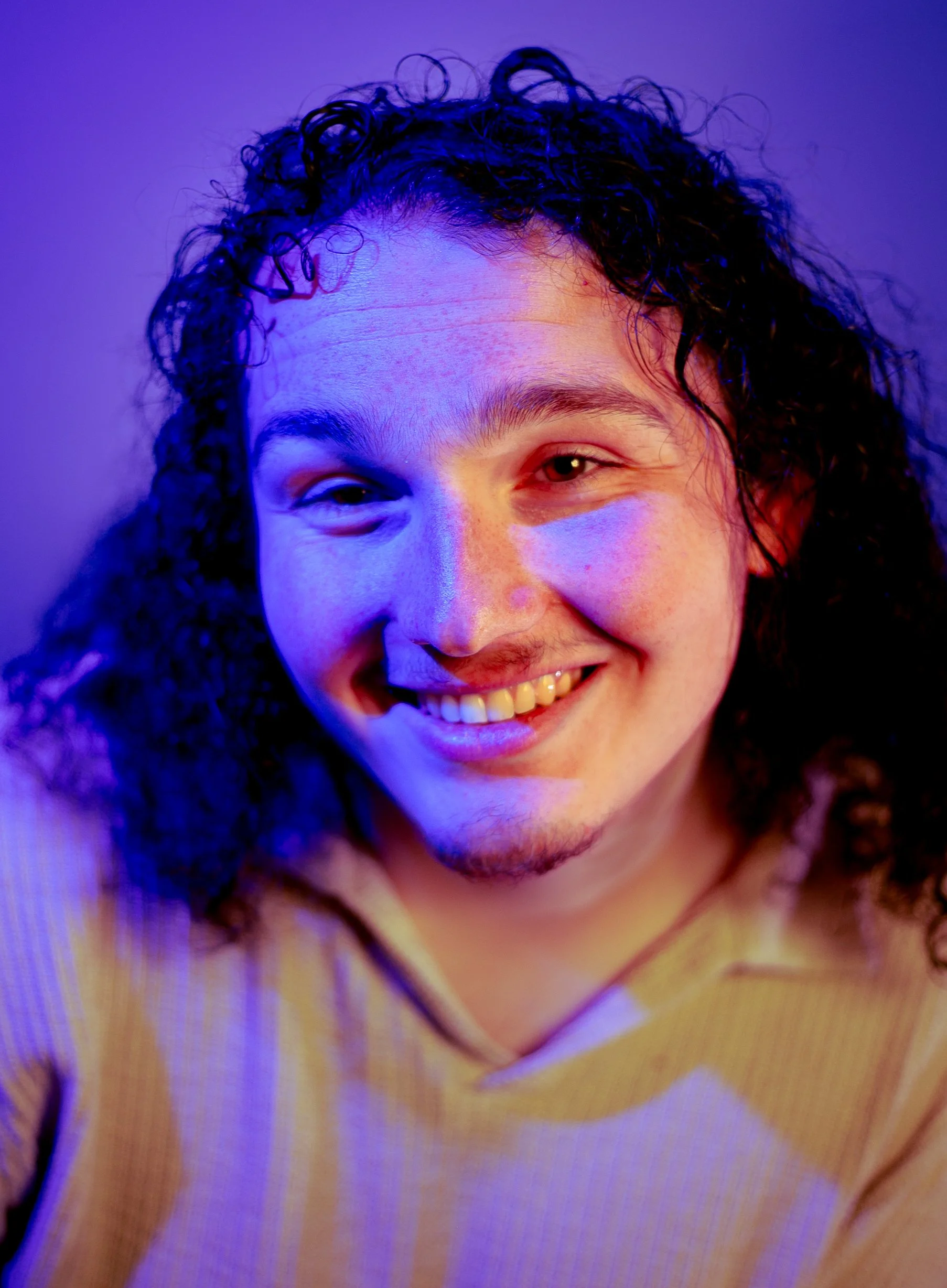Close-up of a smiling person with curly dark hair, wearing a yellow sweater, under colorful purple and pink lighting.
