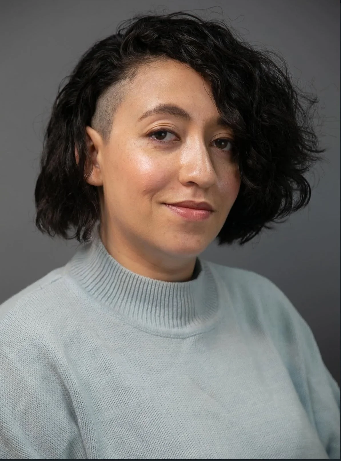 A woman with short, curly black hair, some of it shaved on one side, wearing a light gray turtleneck sweater, smiling softly against a plain gray background.