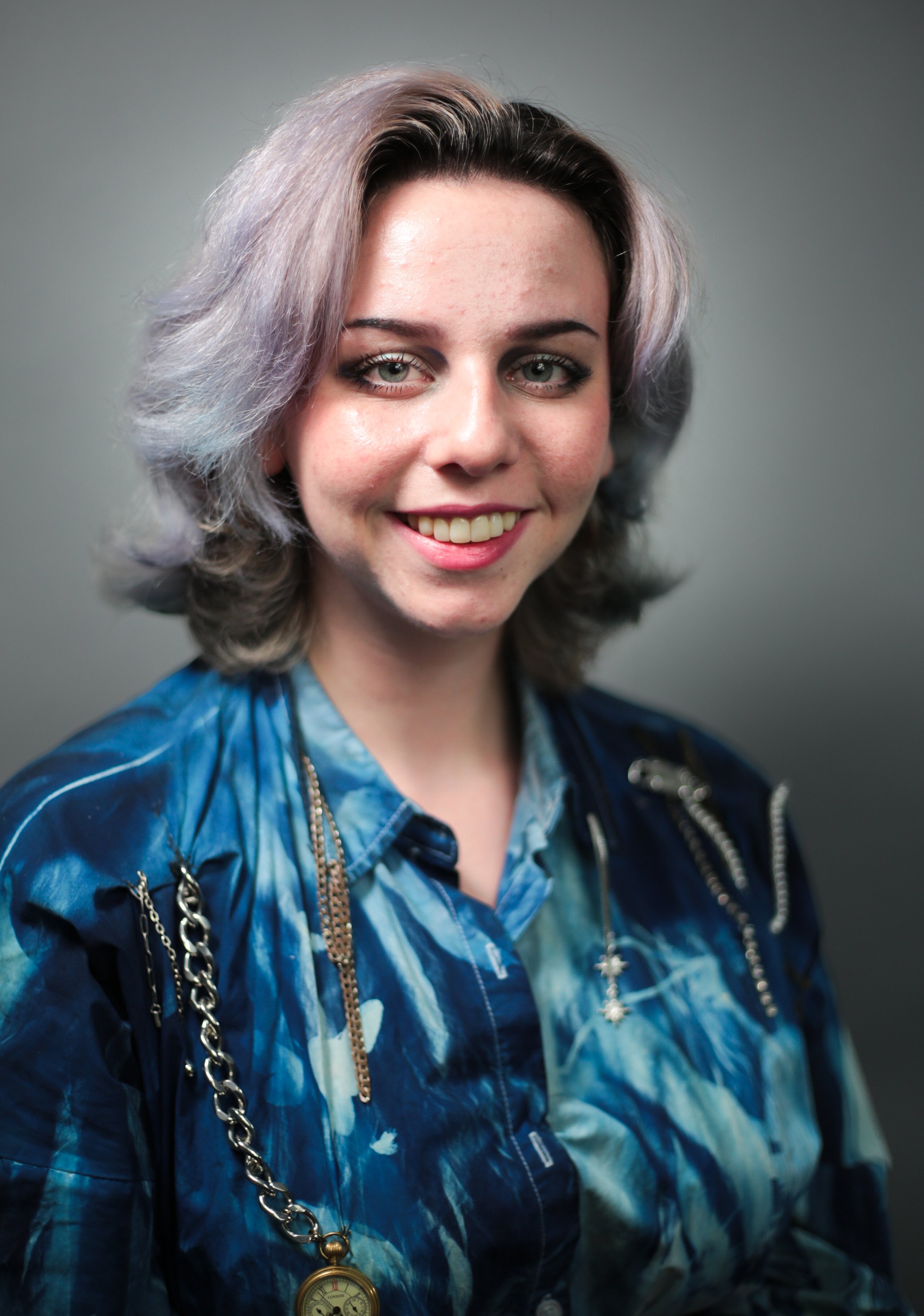 A woman with shoulder-length gray hair smiling at the camera, wearing a blue patterned shirt and jewelry, against a neutral background.