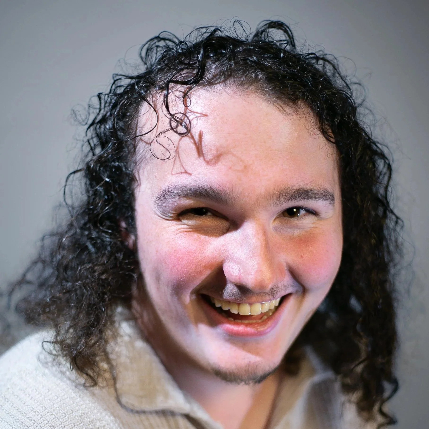 A young man with curly dark hair, light skin, and a cheerful expression, smiling broadly, showing his teeth.