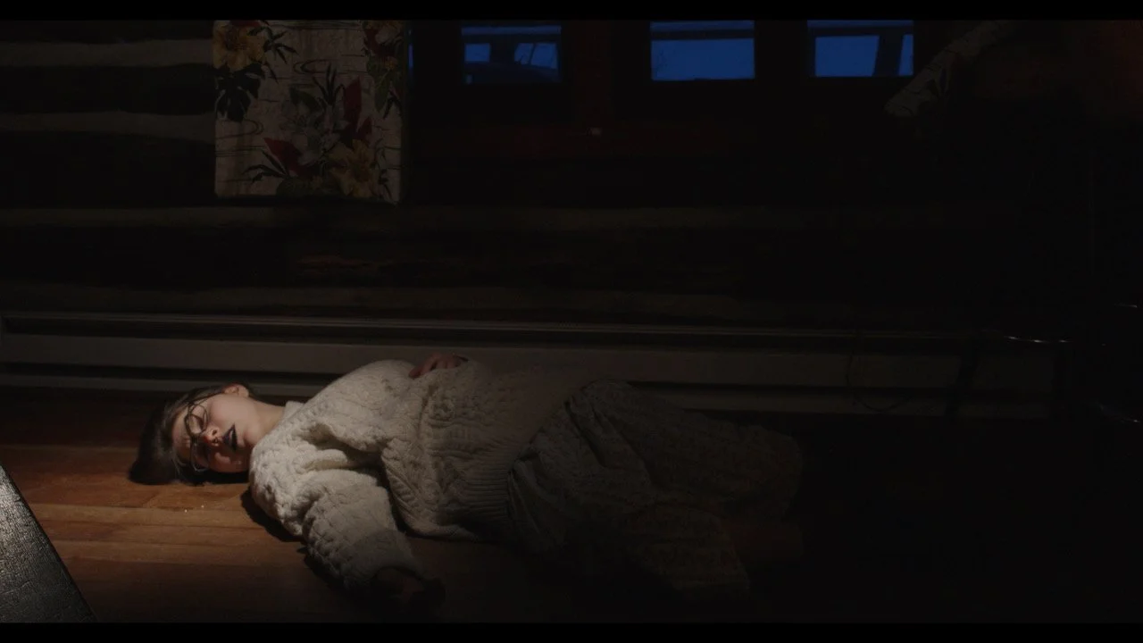 Person lying on the floor, sleeping or resting, in a dimly lit room with curtains and windows in the background.