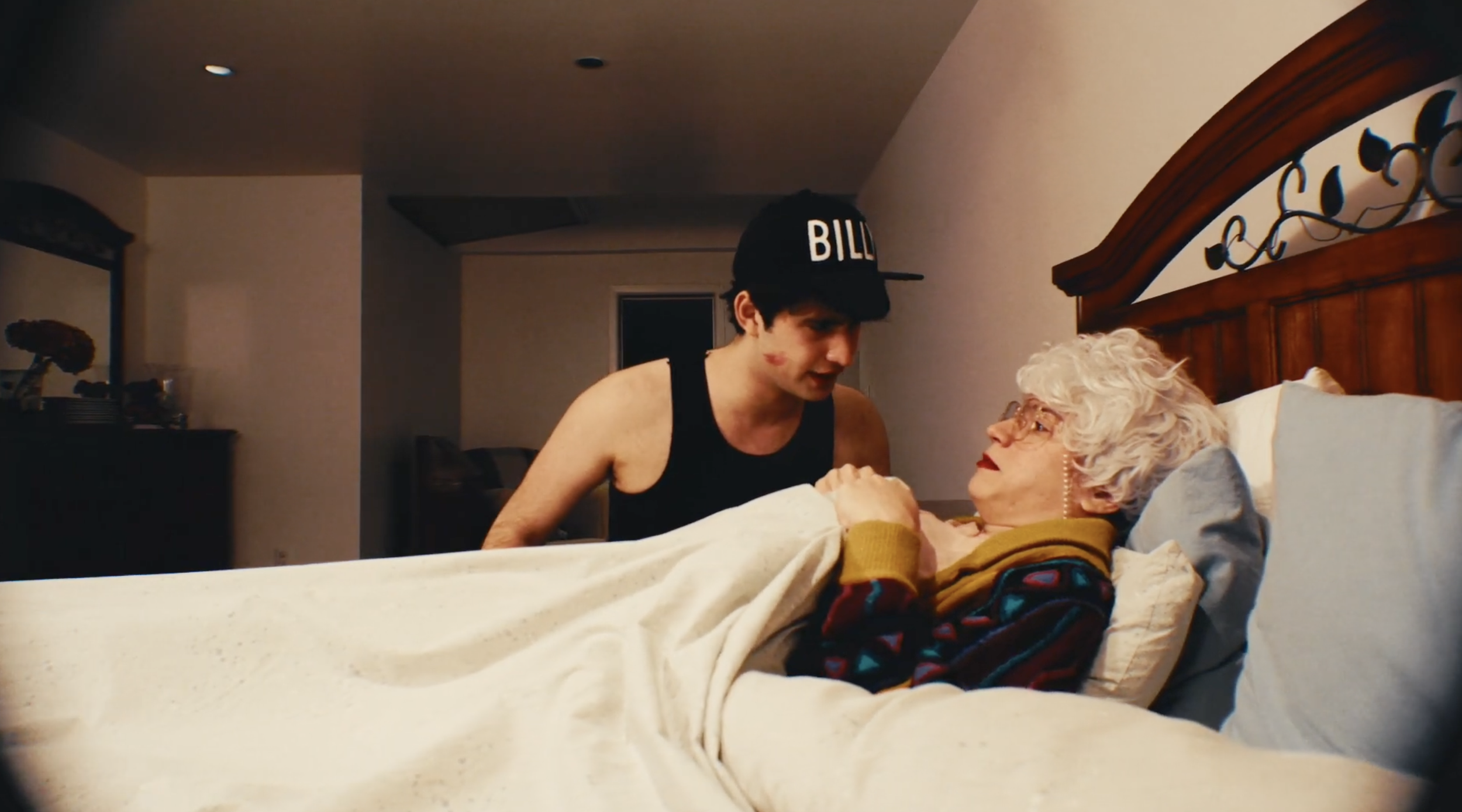 A young man with a black cap labeled 'BILL' leaning over an elderly woman lying in bed, holding her hand and engaging in conversation in a dimly lit bedroom.