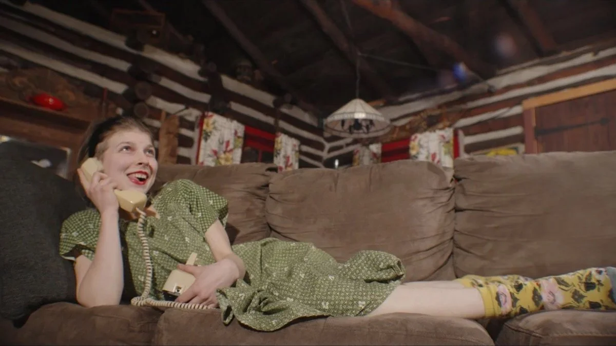 A young girl with dark hair styled in a 1950s fashion, wearing a green patterned dress and yellow floral socks, lying on a brown couch, talking on a beige rotary phone, in a rustic wooden cabin with floral curtains and a stained glass lampshade.