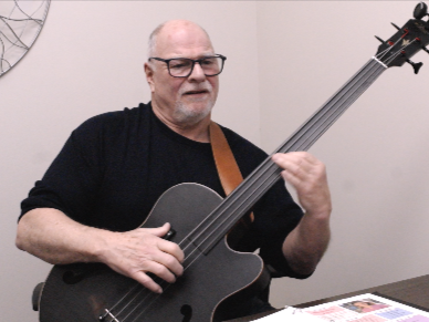 Elderly man playing a black electric cello in an indoor setting.