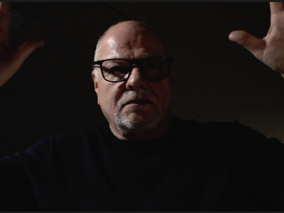A man wearing glasses and a black shirt, with his hands raised, in a dark setting.