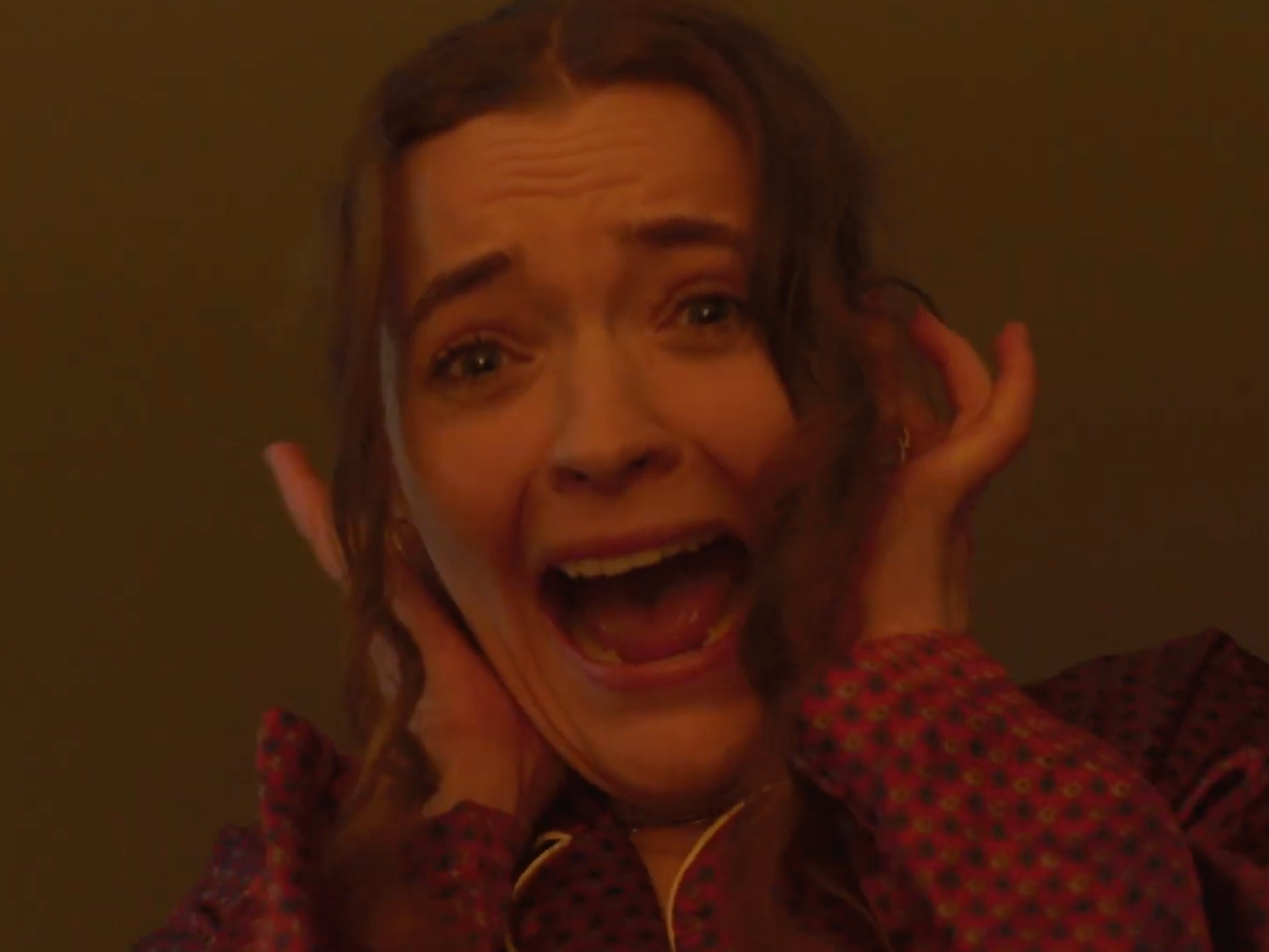 A woman with short, curly hair, wearing a patterned shirt, appears to be in distress or panic, holding her head with her hands, her mouth open, and eyes wide.