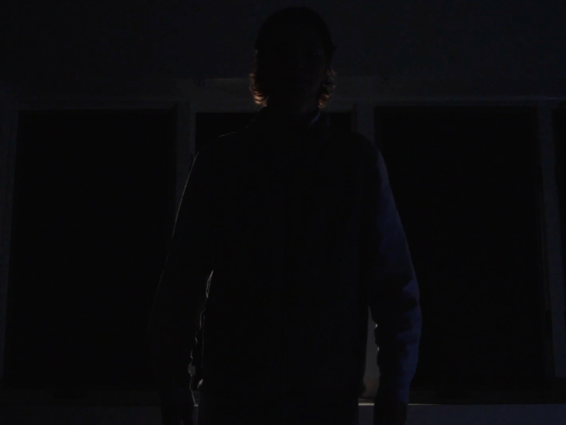 A person standing in a dark room with backlit windows, barely visible due to low lighting.