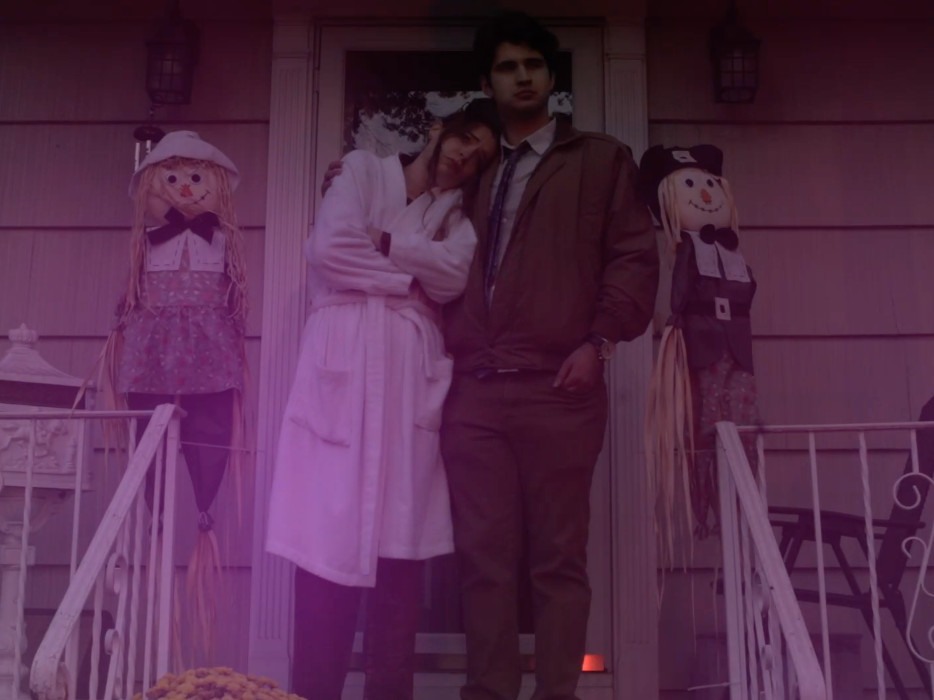 Two young men standing on a porch with Halloween decorations, including two scarecrow figures on either side of the door.
