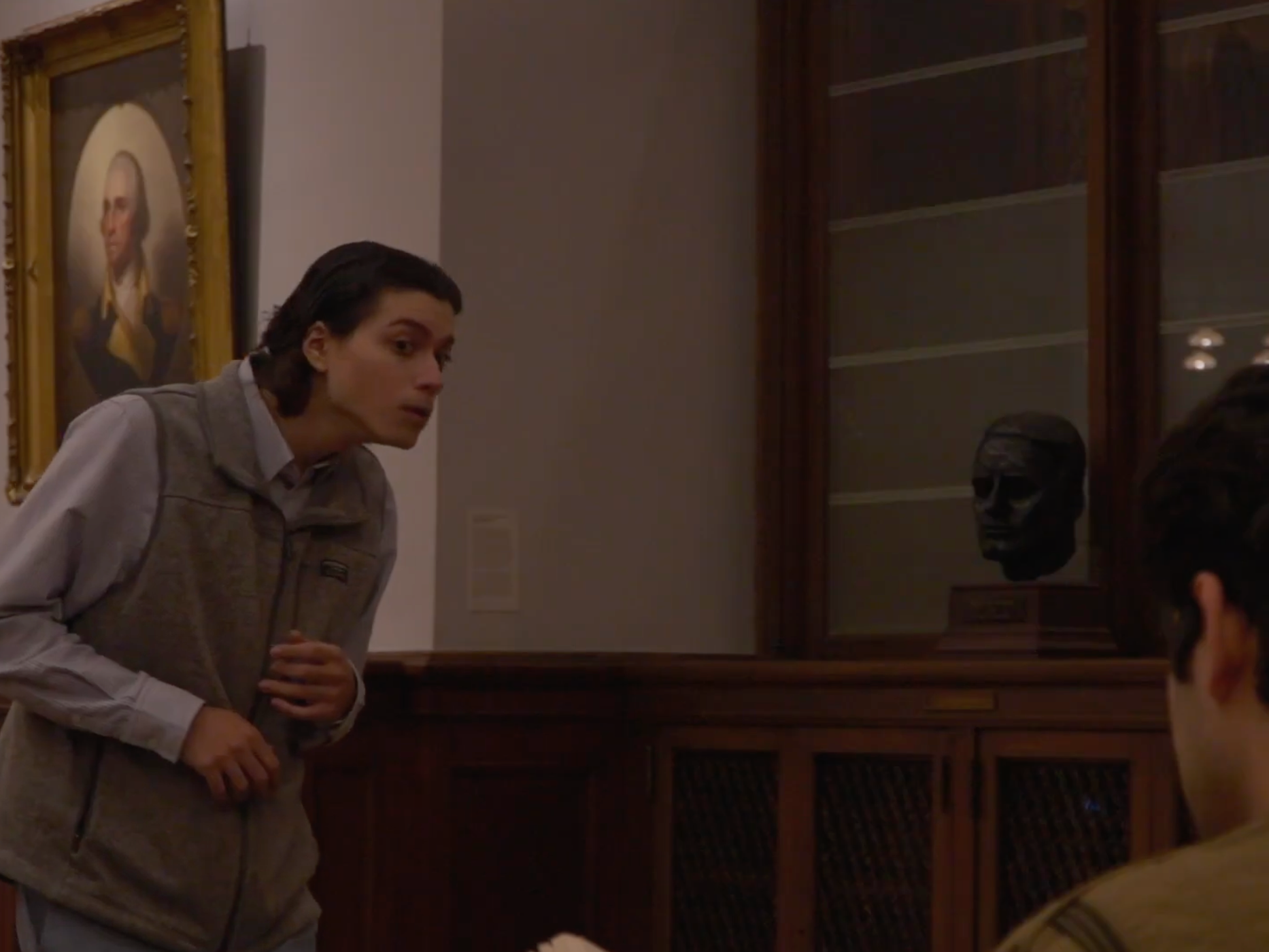A woman with short dark hair leaning forward and speaking to a man whose back is to the camera in a museum or gallery, with a portrait of George Washington and a display case with a dark skull-shaped object behind them.