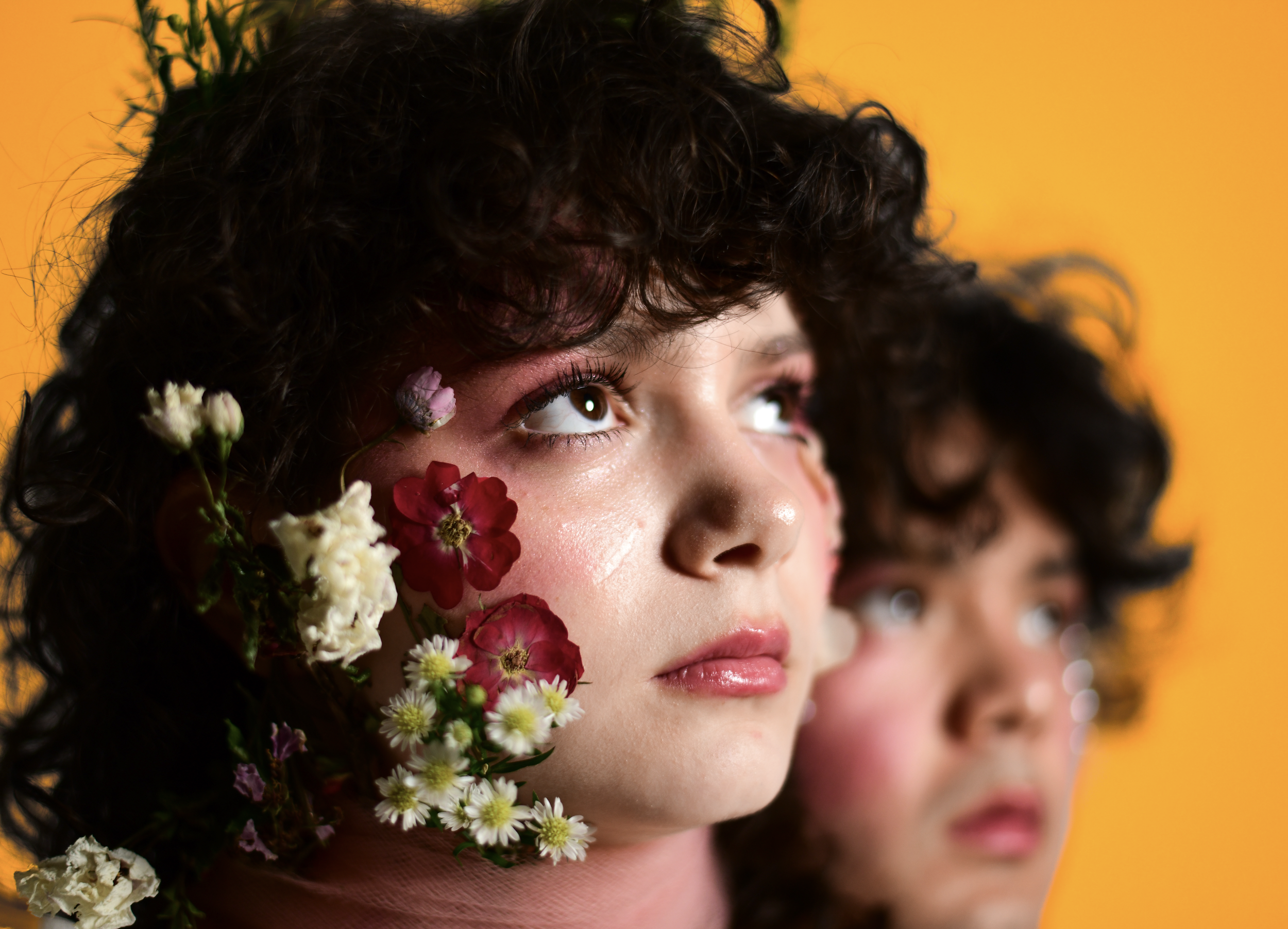 Close-up of a woman with curly dark hair, adorned with flowers on her face, looking upward against a bright yellow background.