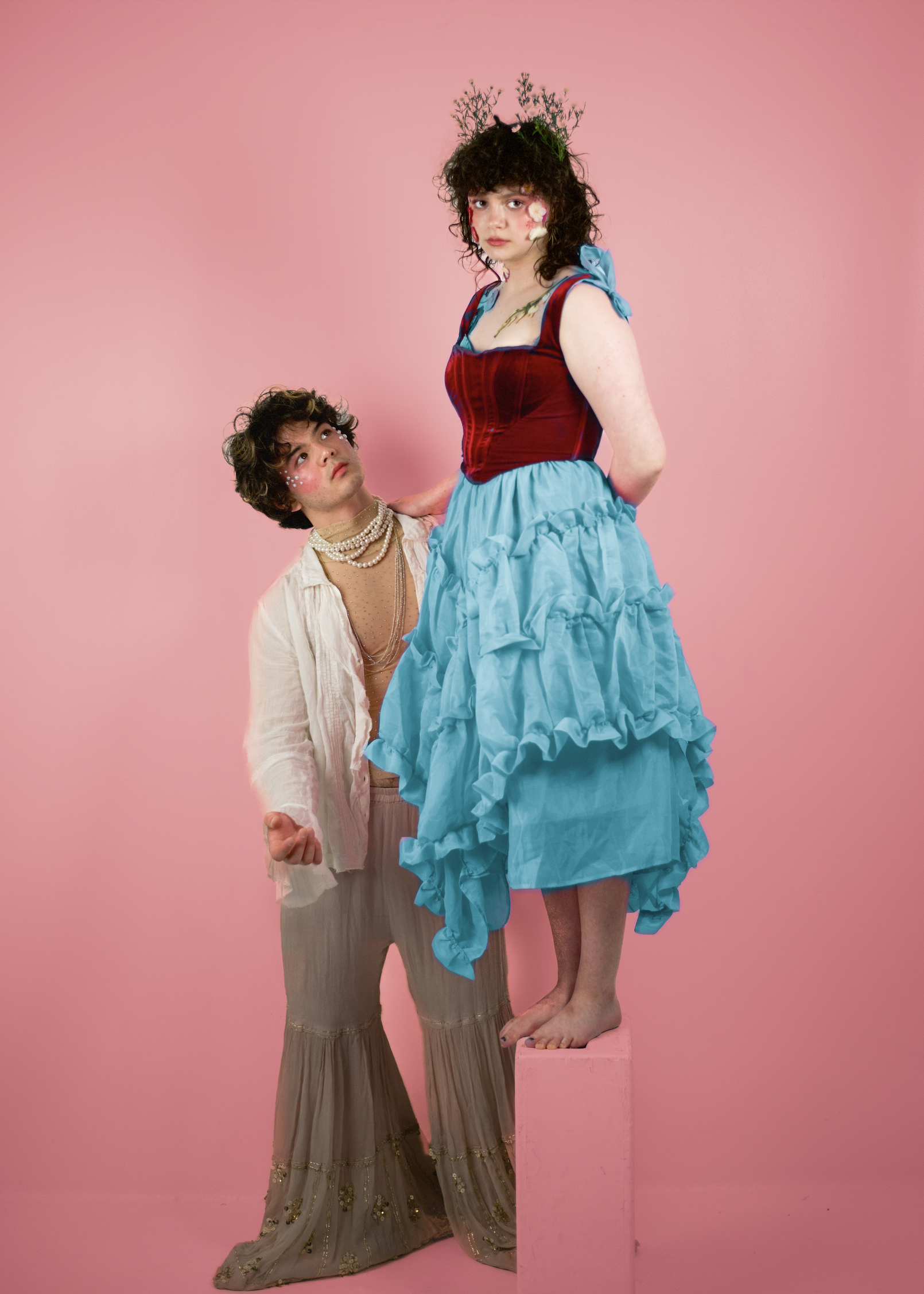 Two individuals dressed in colorful, whimsical costumes standing against a pink background. One person is standing on a pink pedestal, wearing a blue ruffled skirt, a red corset, and a crown of twigs, with flowers on their face and shoulder. The other person is standing beside them, looking up, wearing beige bell-bottom pants with gold embellishments, a cream-colored shirt, layered pearl necklaces, and makeup with white dots around the eyes