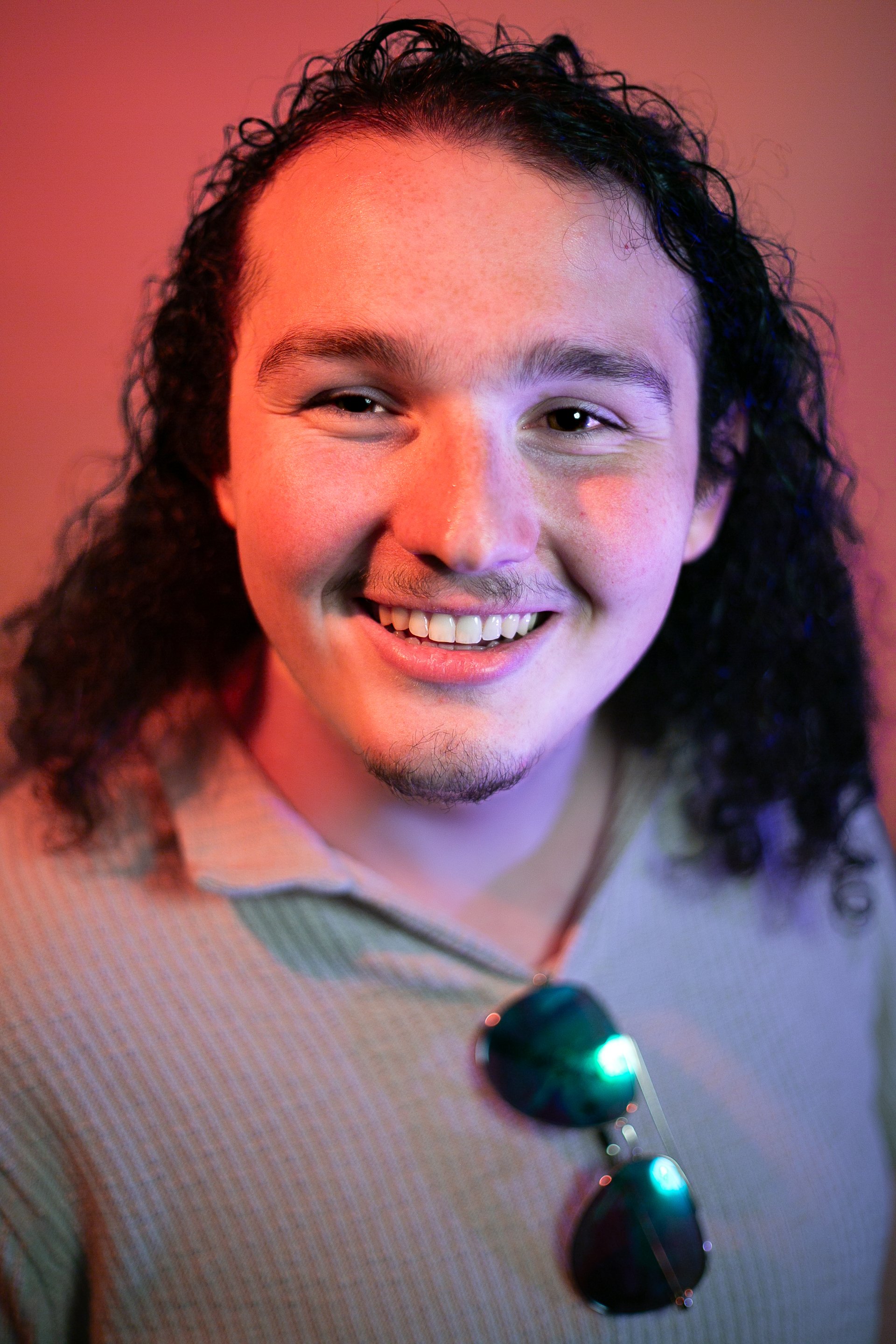 A young man with curly dark hair, a light mustache, and goatee, smiling, wearing a beige shirt with sunglasses hanging from the collar, against a colorful pink and purple background.