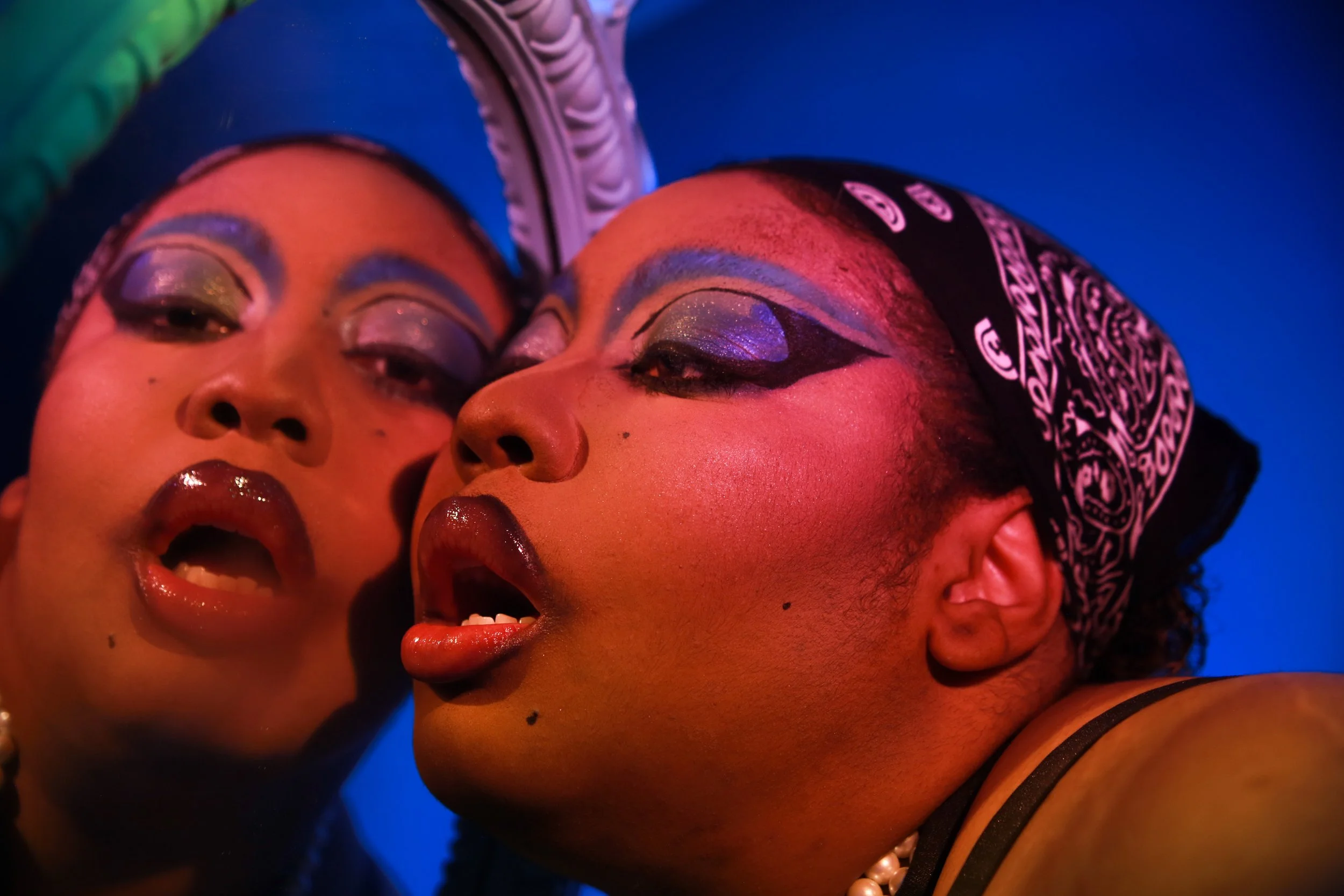 Two women with dramatic, colorful makeup, posing closely together with faces near each other against a vibrant background.