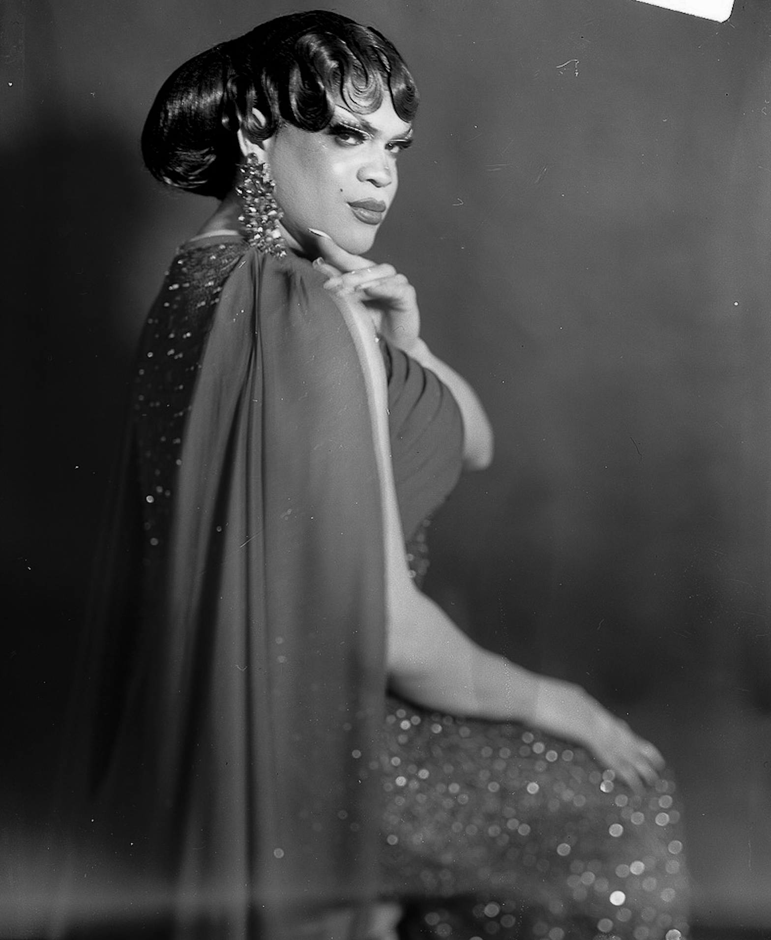Black and white film photograph of a woman with styled hair, wearing a glamorous dress and earrings, posing with one hand resting on her chin against a plain background.