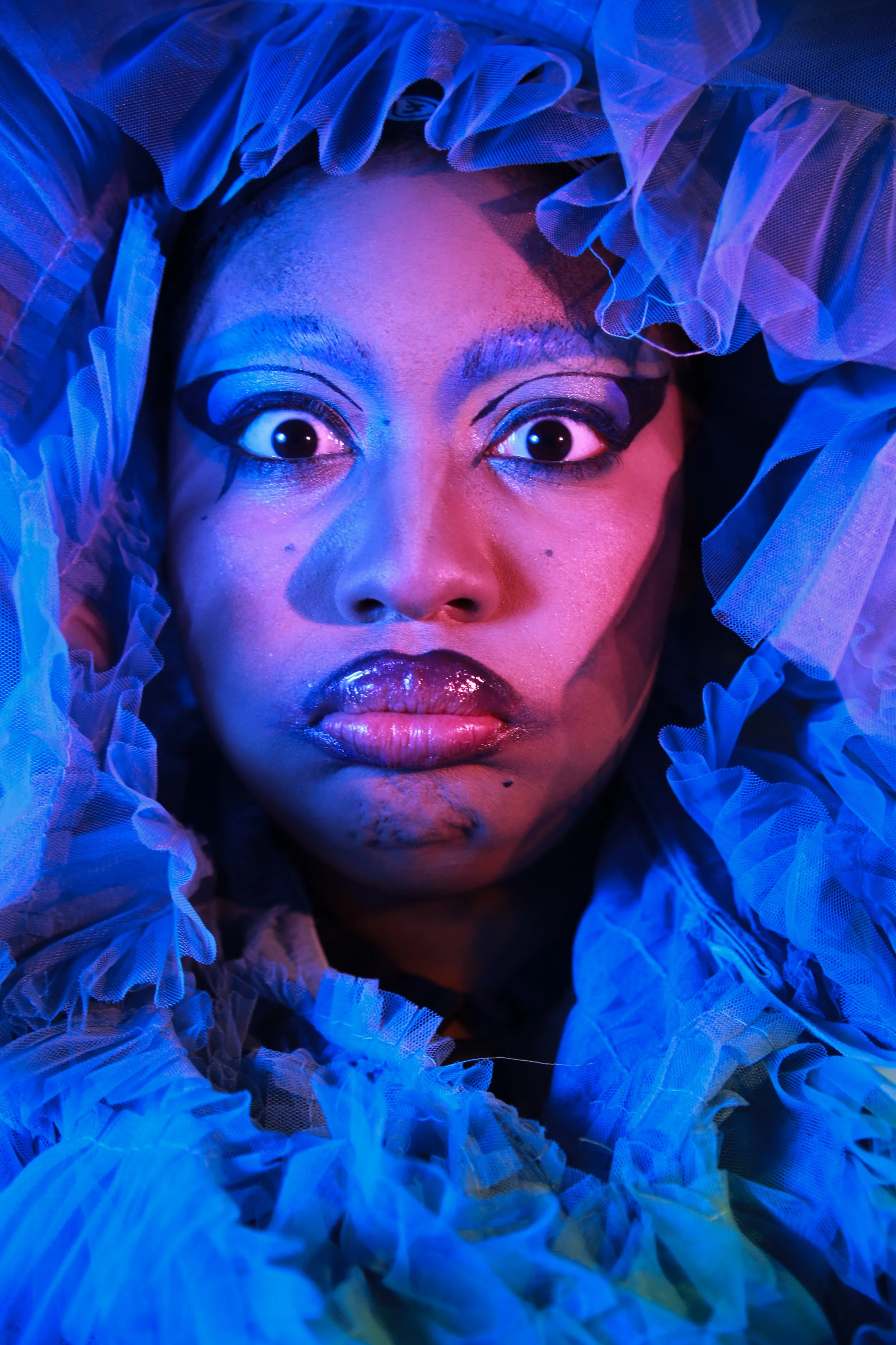 Close-up of a woman's face surrounded by ruffled, blue-tinted fabric with dramatic makeup and glossy lips, under colorful lighting.