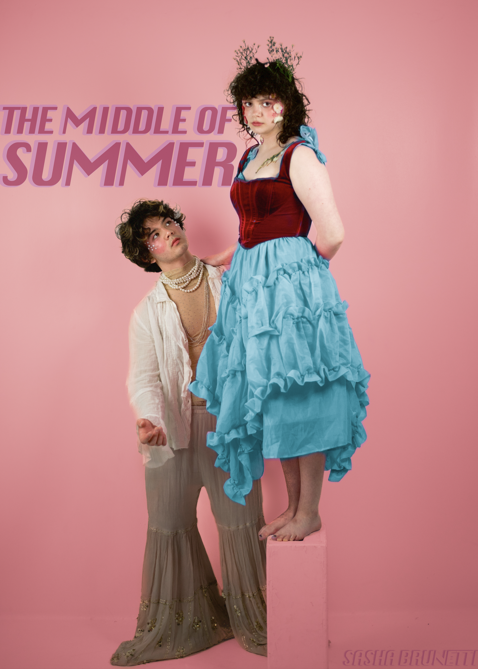 Two young people dressed in vintage clothing, one standing on a pink pedestal and the other standing next to them, in front of a pink background with purple text that reads 'The Middle of Summer.' The person standing on the pedestal has curly dark hair, a red and blue dress, and floral face makeup. The other person has short curly dark hair, layered necklaces, and wears a beige and white outfit with embroidered details.