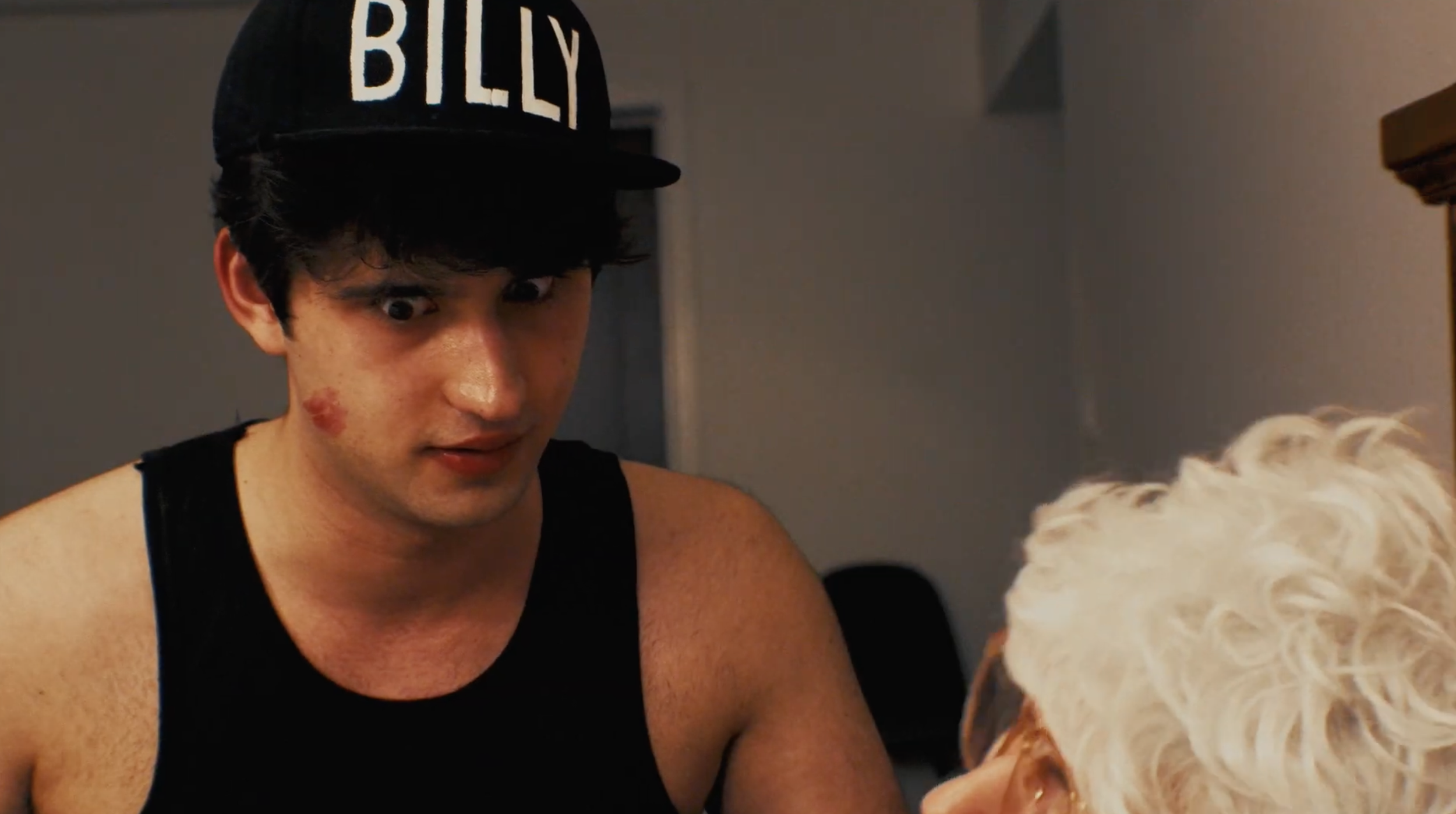 Young man wearing a black cap with 'BILLY' on it, looking at a fluffy white dog.
