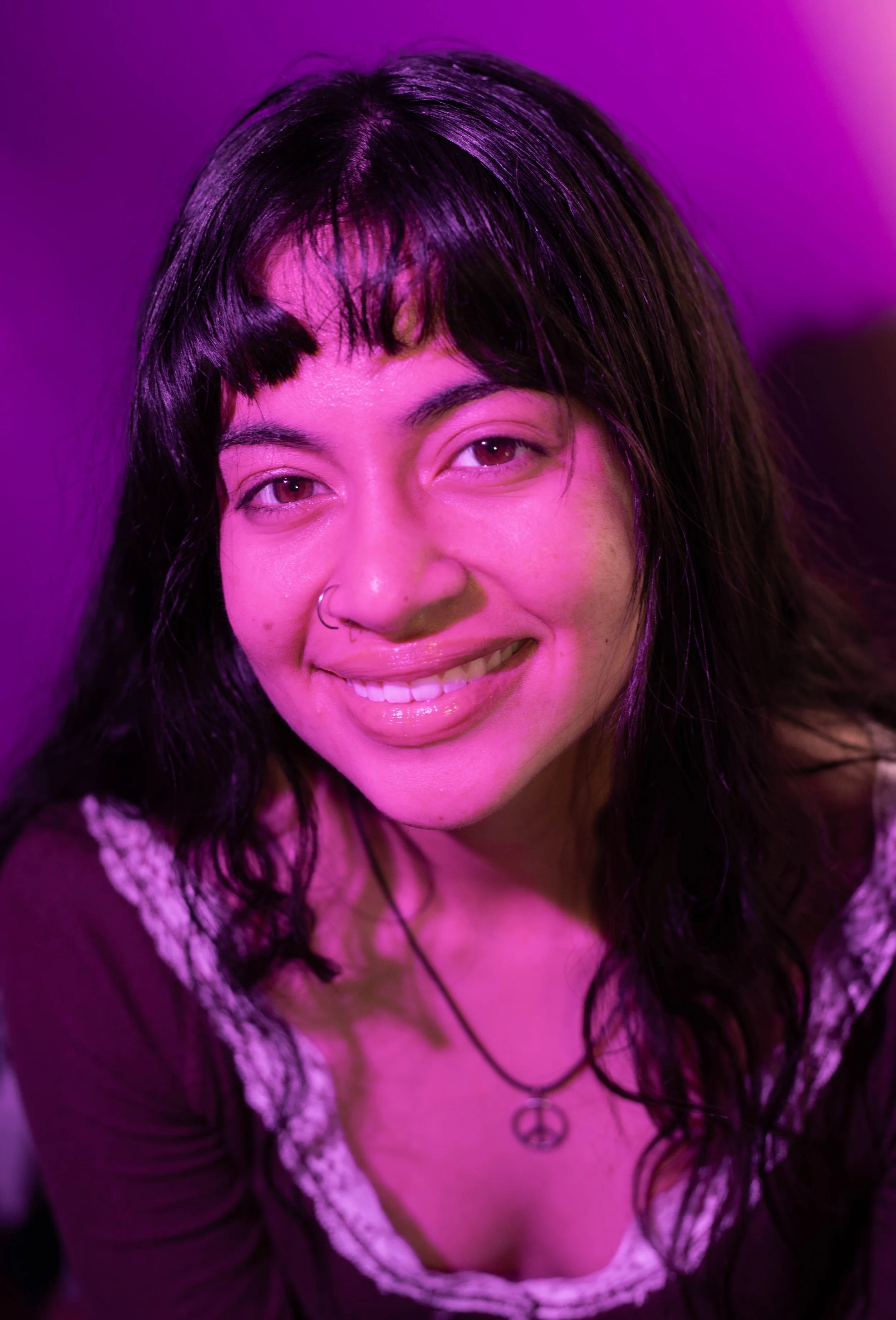 Close-up portrait of a woman with dark, wavy hair, wearing a peace sign necklace and smiling, illuminated by pink and purple lighting.
