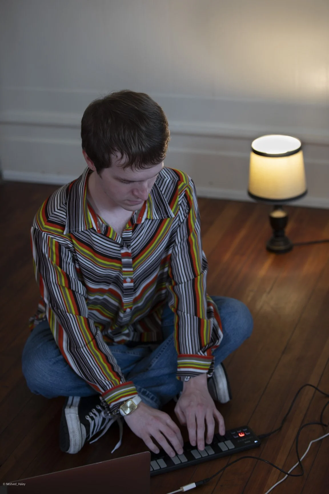 Young man sitting cross-legged on the floor, playing a small electronic musical keyboard with a laptop nearby, in a room with wooden floor and a lamp in the background.