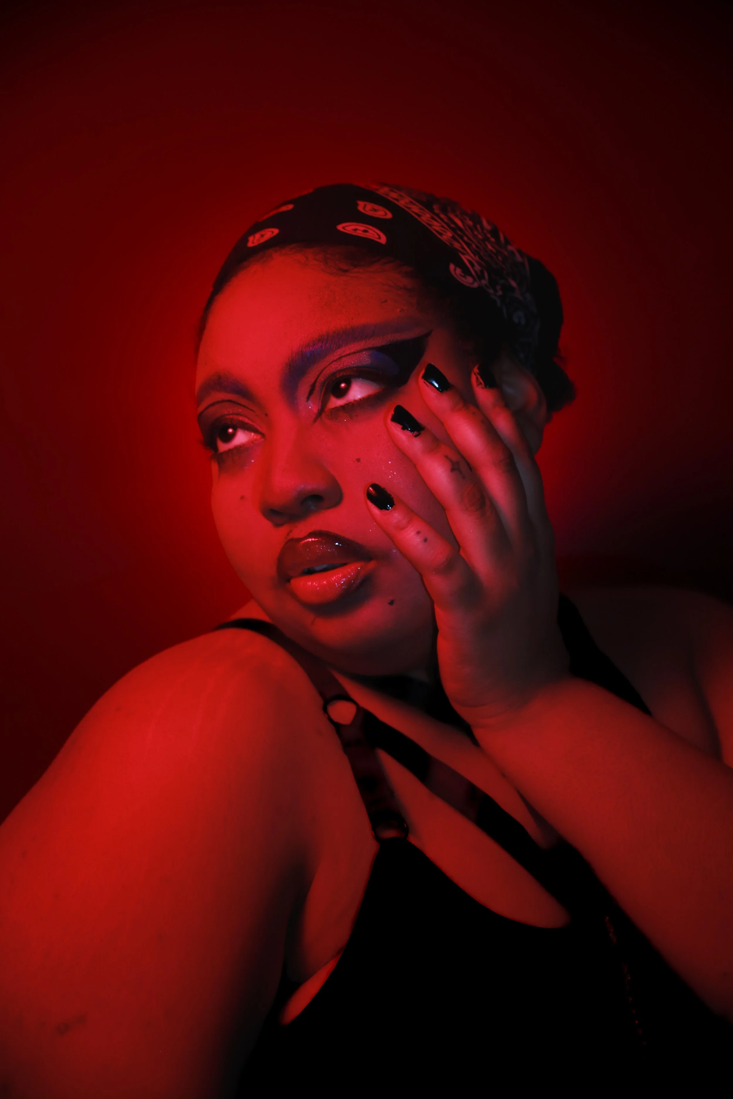 A woman with dark makeup and a bandana on her head, touching her face with her hand, against a red background.