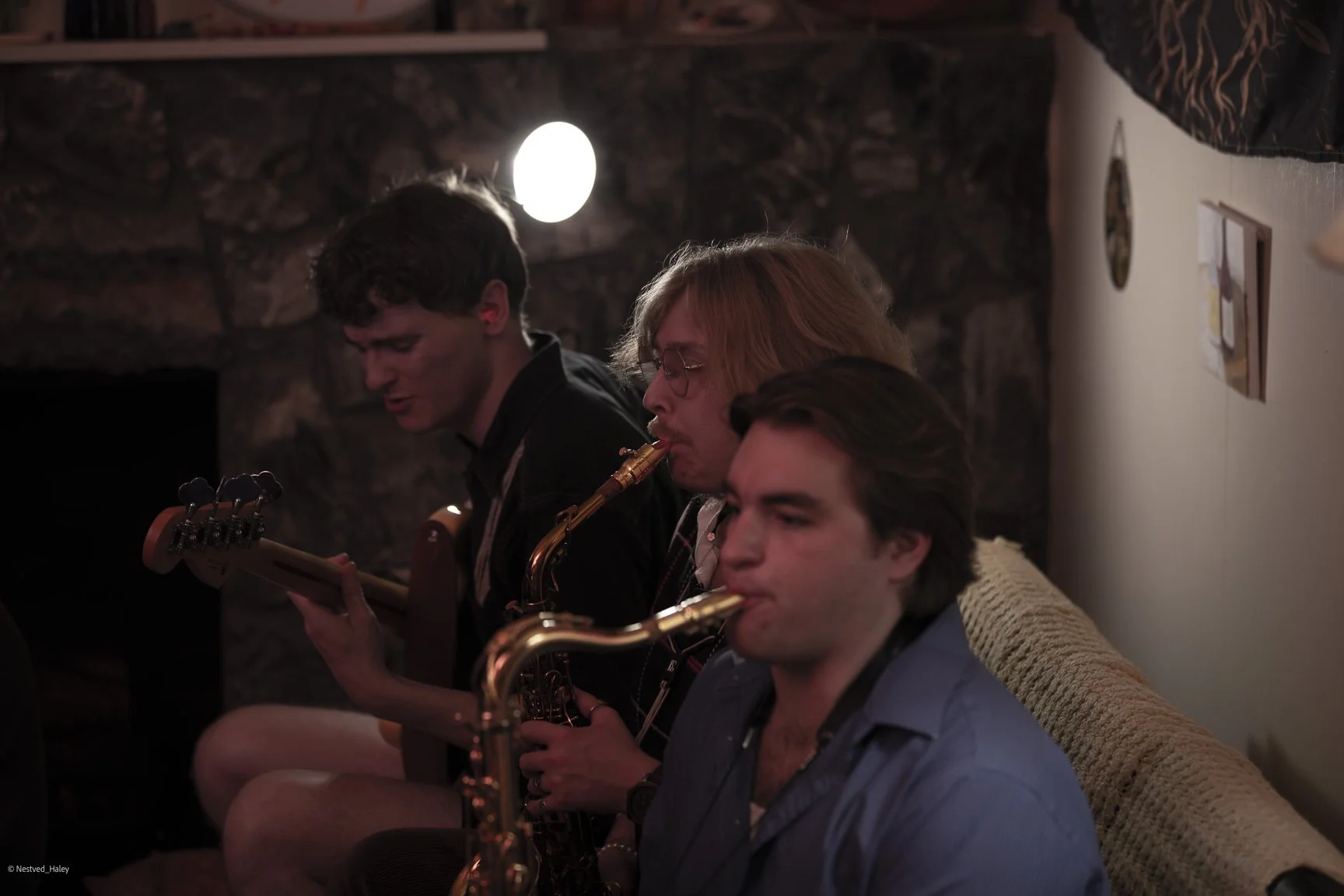 Three men sitting closely together playing musical instruments in a dimly lit room with a white wall background.
