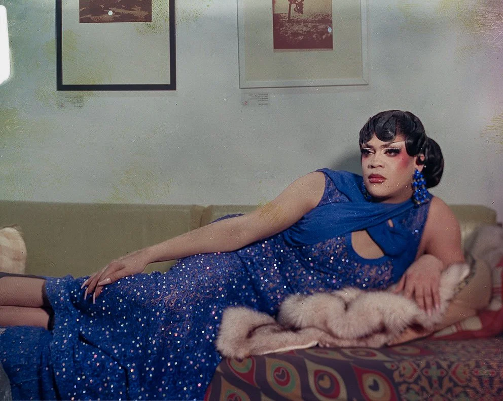 A person dressed in a glamorous blue dress with sequins, lying on a couch with a fur stole, in a room with framed pictures on the wall.