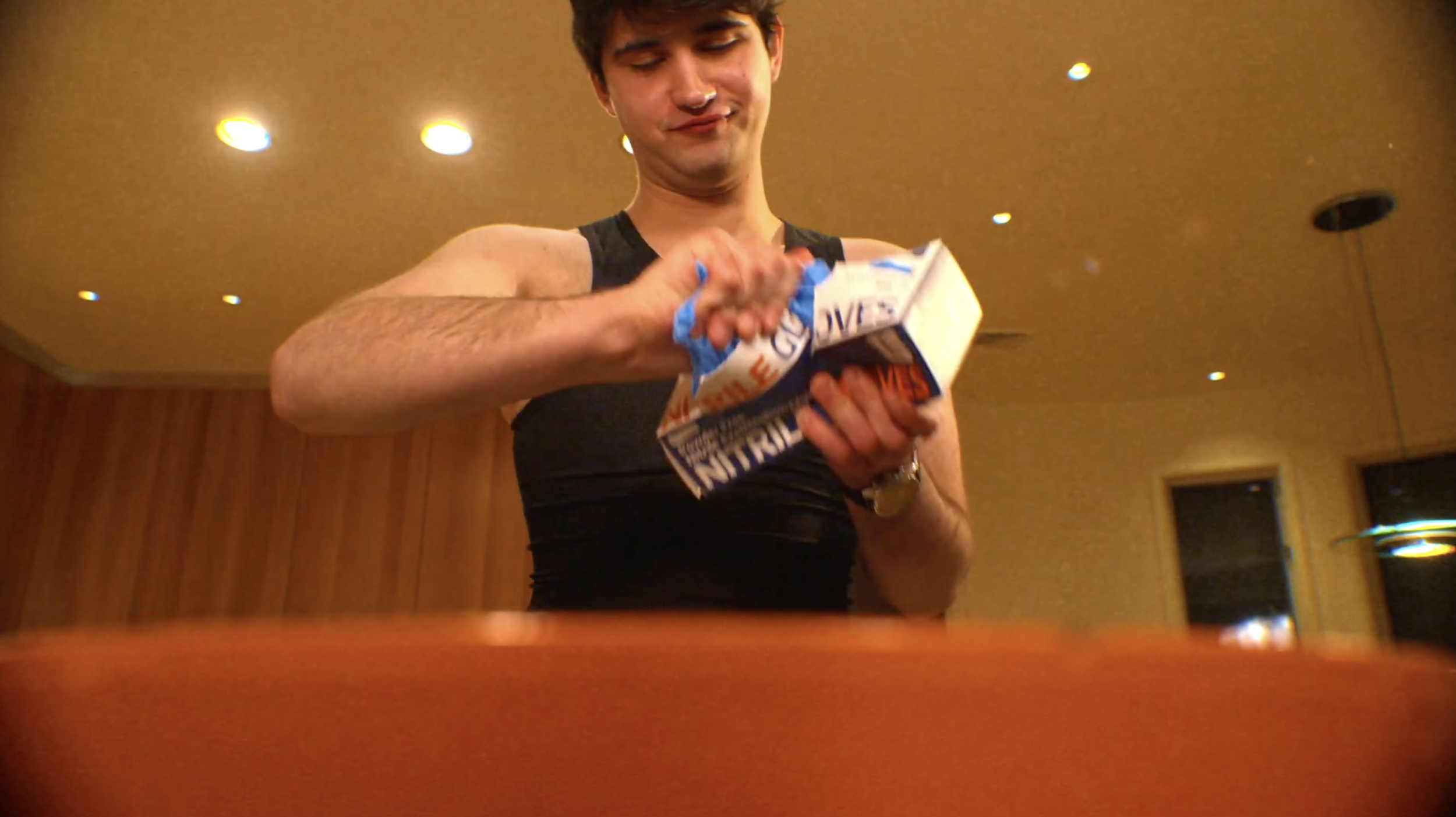 A young man with dark hair wearing a black sleeveless shirt is opening a box of Little Debbie Swiss Rolls in a room with wooden paneling and hanging ceiling lights.