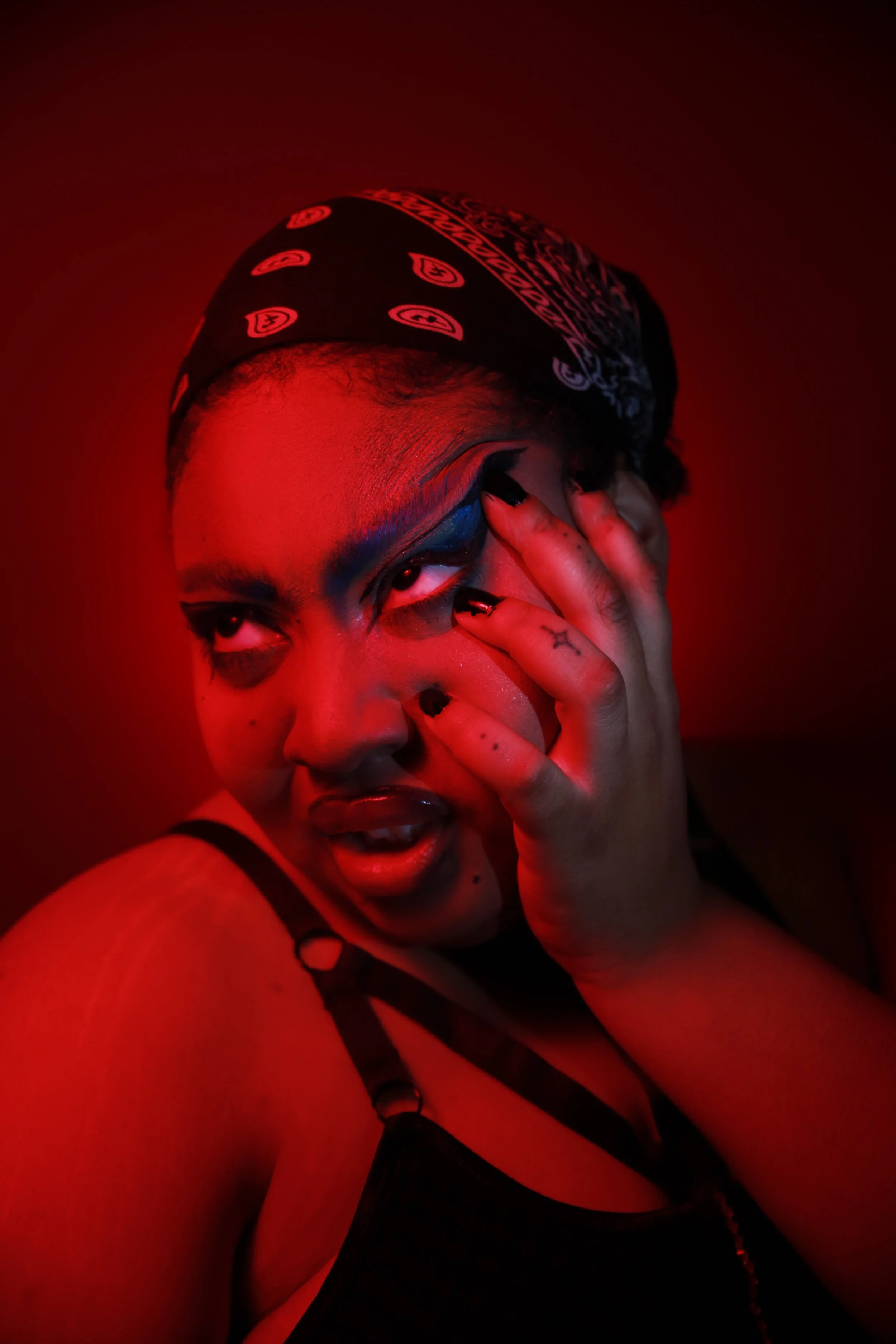 A person with dark makeup, a bandana on their head, and black nail polish is posing with their hand on their face, with a red lighting effect.